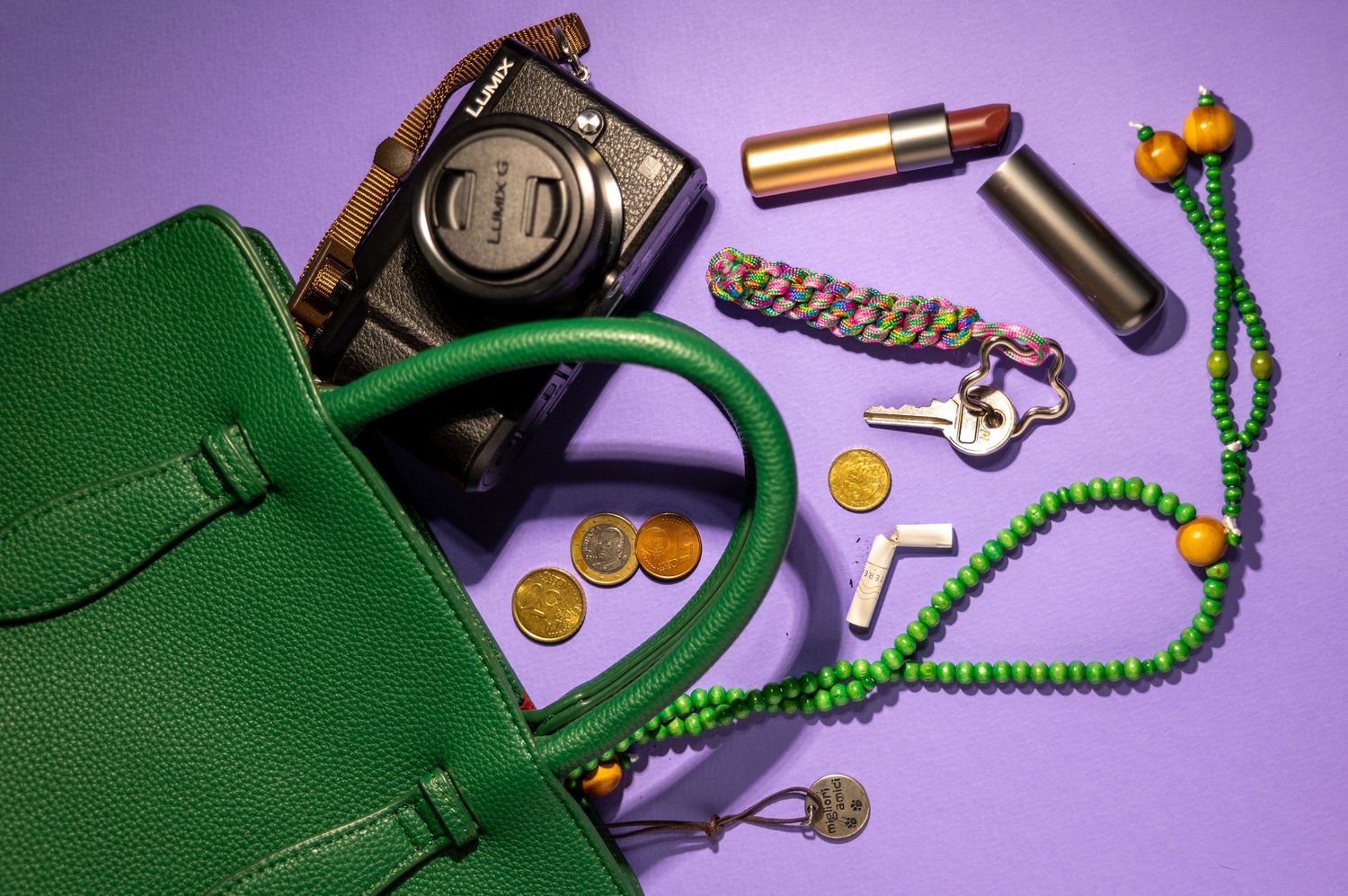 A green handbag spills open to reveal a camera, makeup, coins, keys and colorful beaded accessories against a purple background.