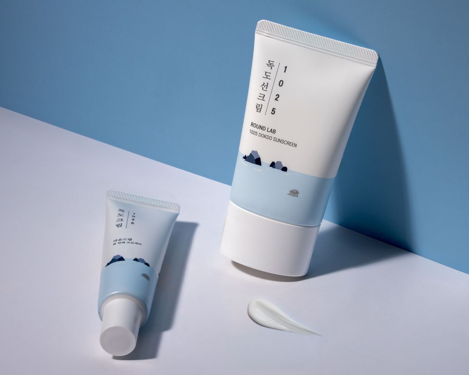 Two minimalist white skincare tubes with Korean text against a blue and white geometric background in studio lighting.