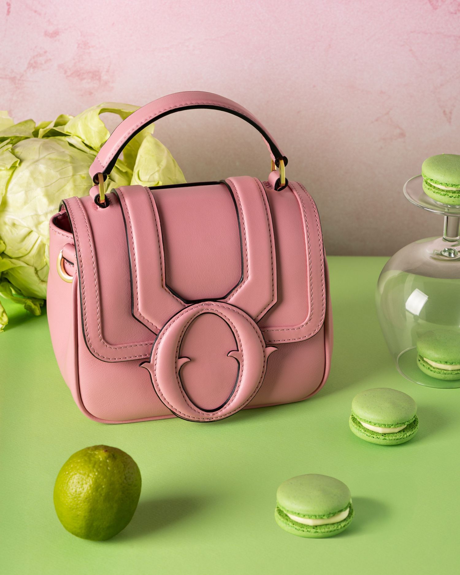 Pink leather handbag styled with green macarons and lime on a green surface against a pink textured background.