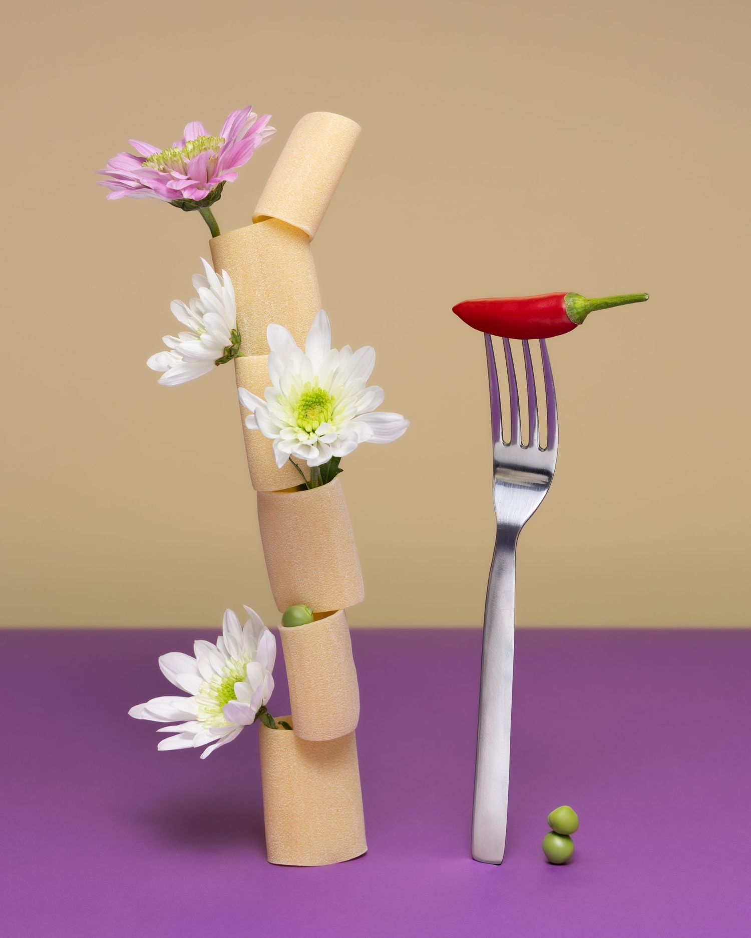Pasta cylinders with colorful flowers intertwined, alongside a fork with a red chili pepper on purple surface.