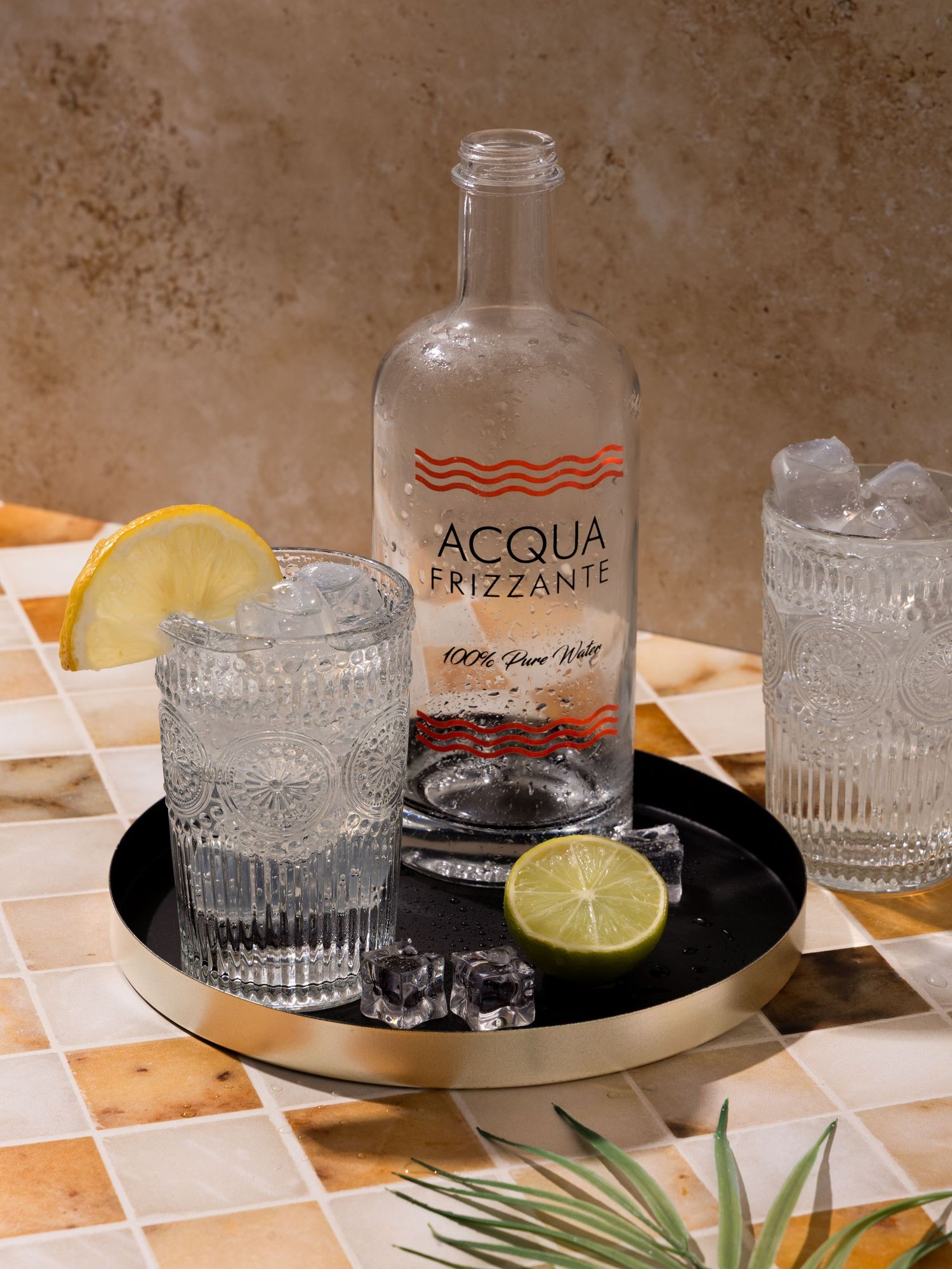 A bottle of Acqua Frizzante sparkling water with glasses, citrus slices, and ice on a patterned surface.