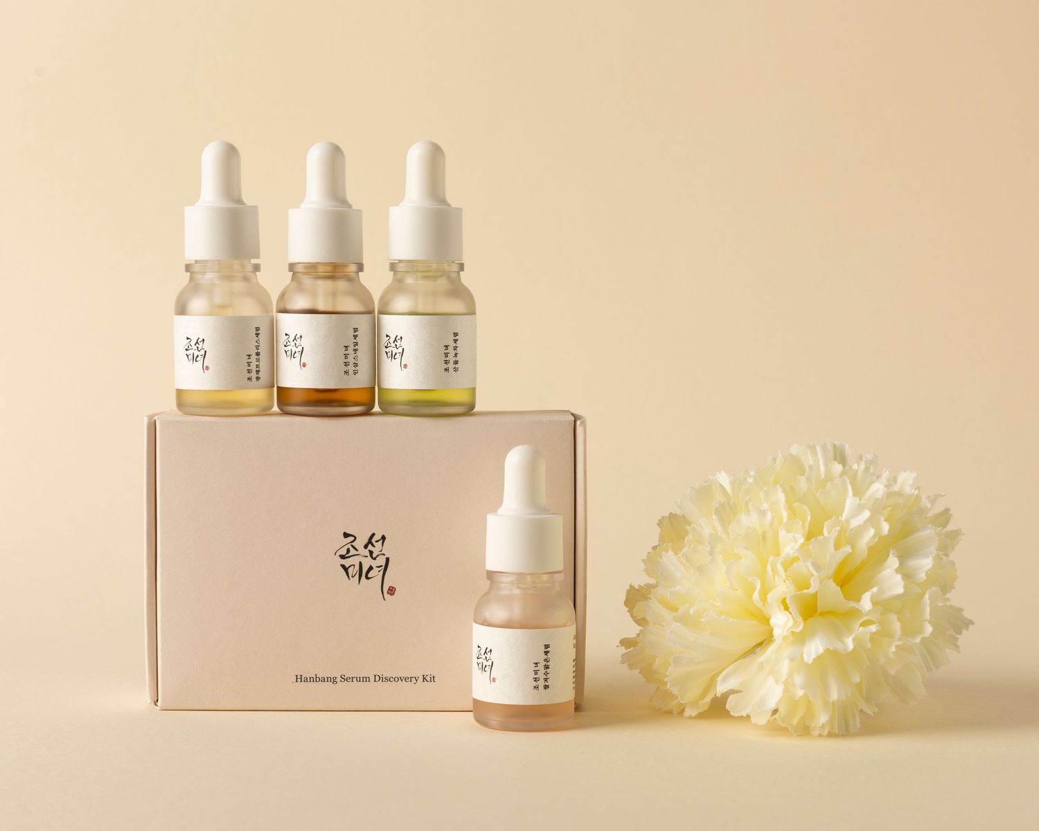 Minimalist arrangement of white dropper bottles and pale yellow carnation flowers on beige background.