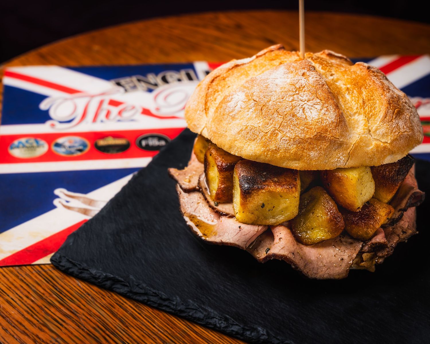 Gourmet beef burger with roasted potatoes served on black slate with British Union Jack flag in background.