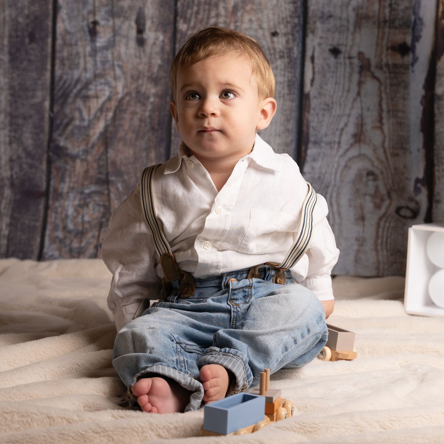 Small child wearing white shirt and denim overalls sits on light carpet with wooden toys beside them.