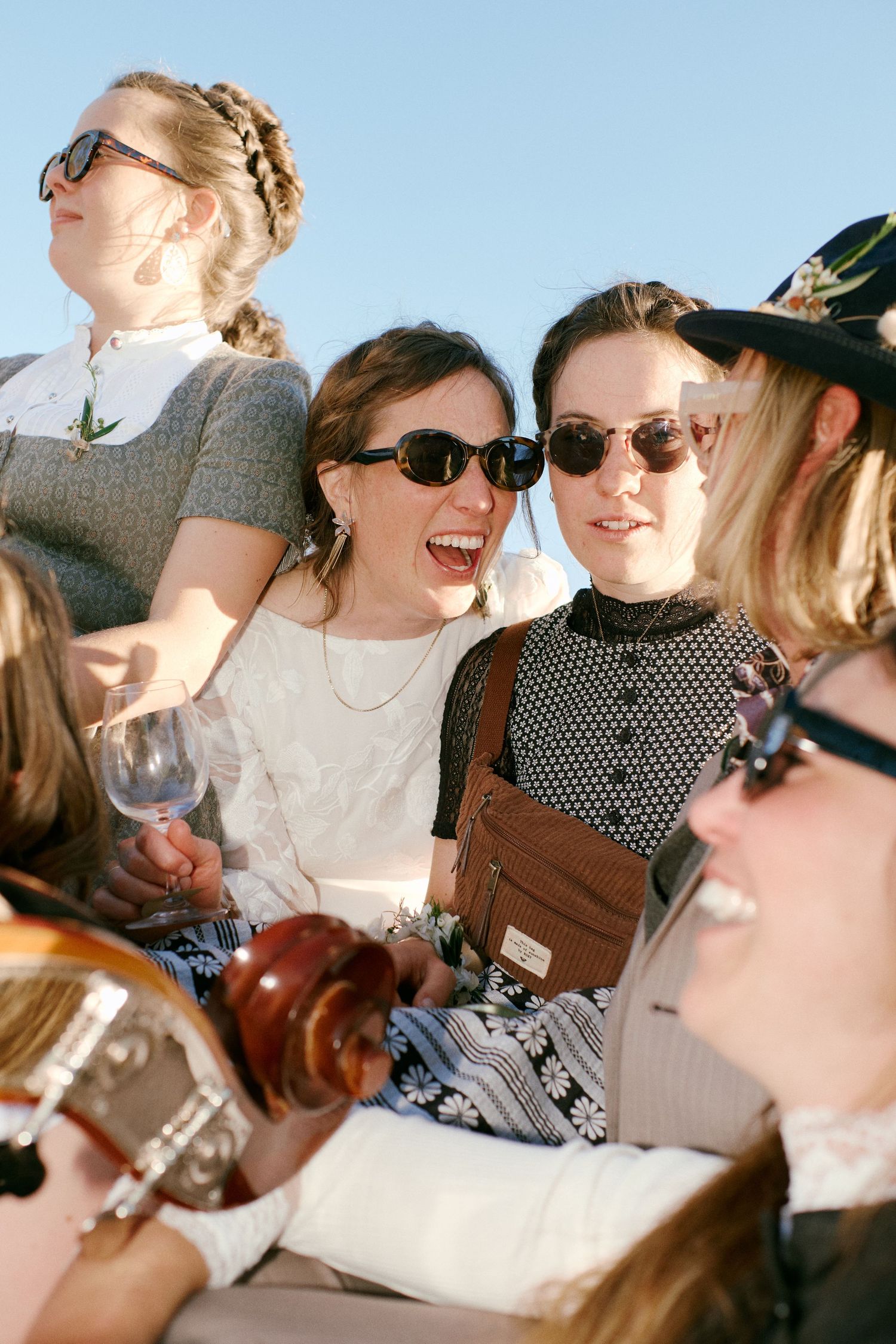 Group of young women laughing and socializing outdoors, wearing sunglasses, one holding a wine glass.