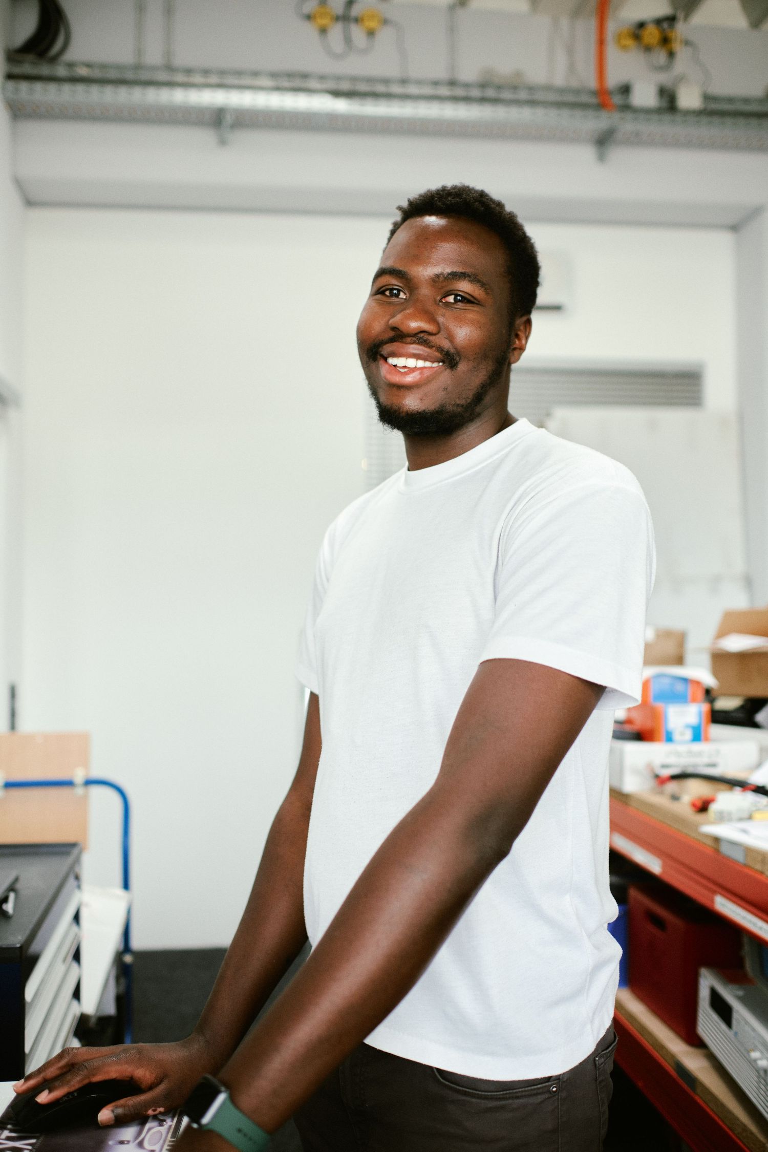 Smiling young man in white t-shirt standing at a workbench in an industrial workspace.