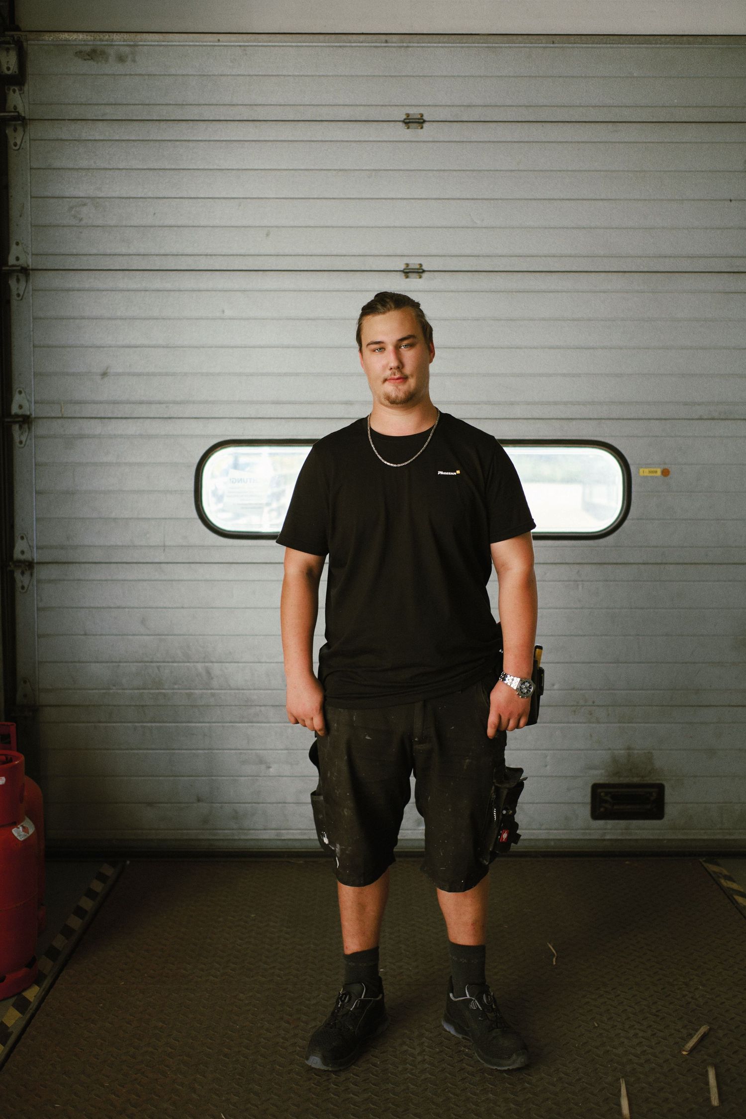 Young mechanic in black t-shirt and cargo shorts stands confidently in front of a garage door.