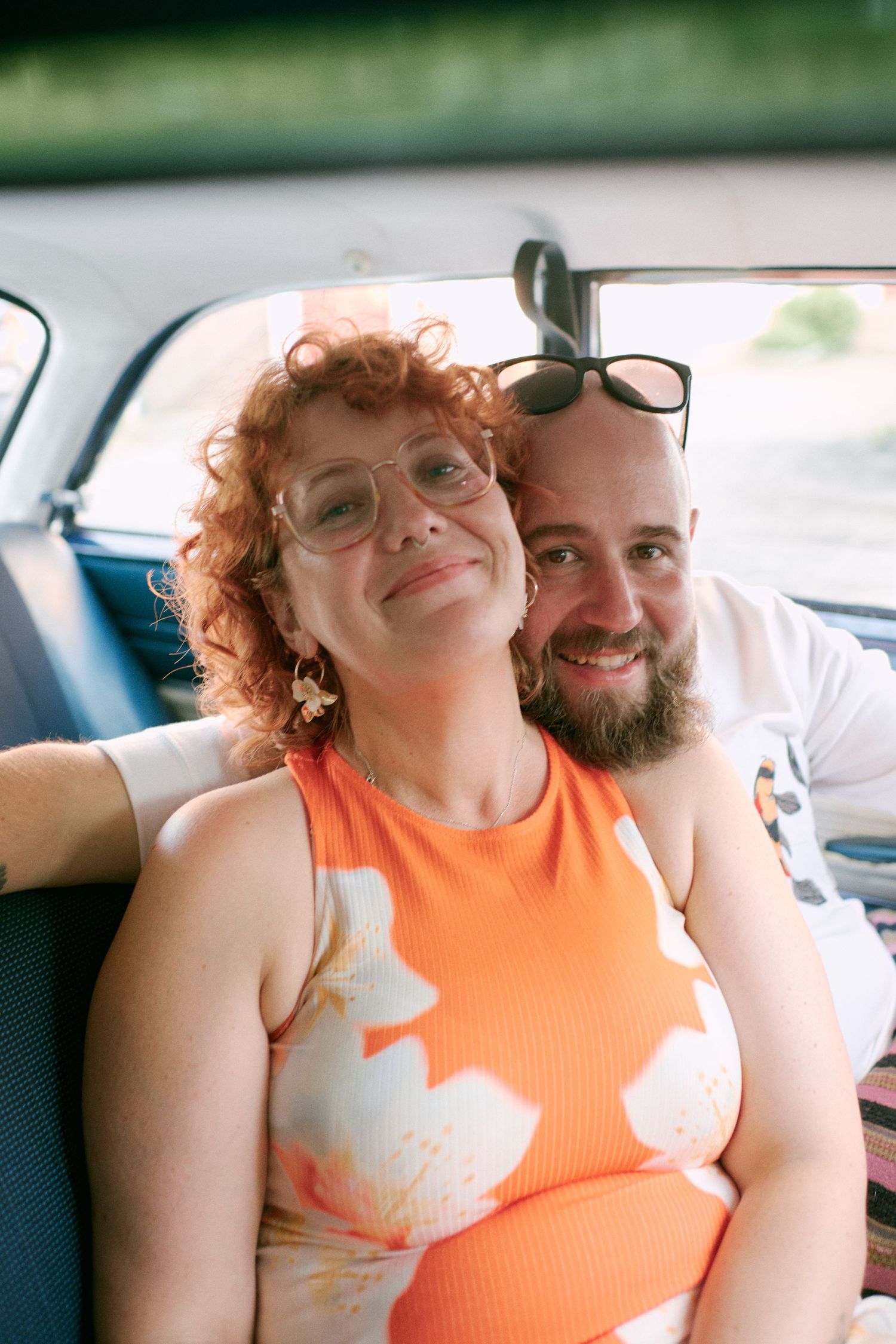 A smiling couple sits together in a car backseat, woman in orange floral dress with curly red hair, man with beard and sunglasses.