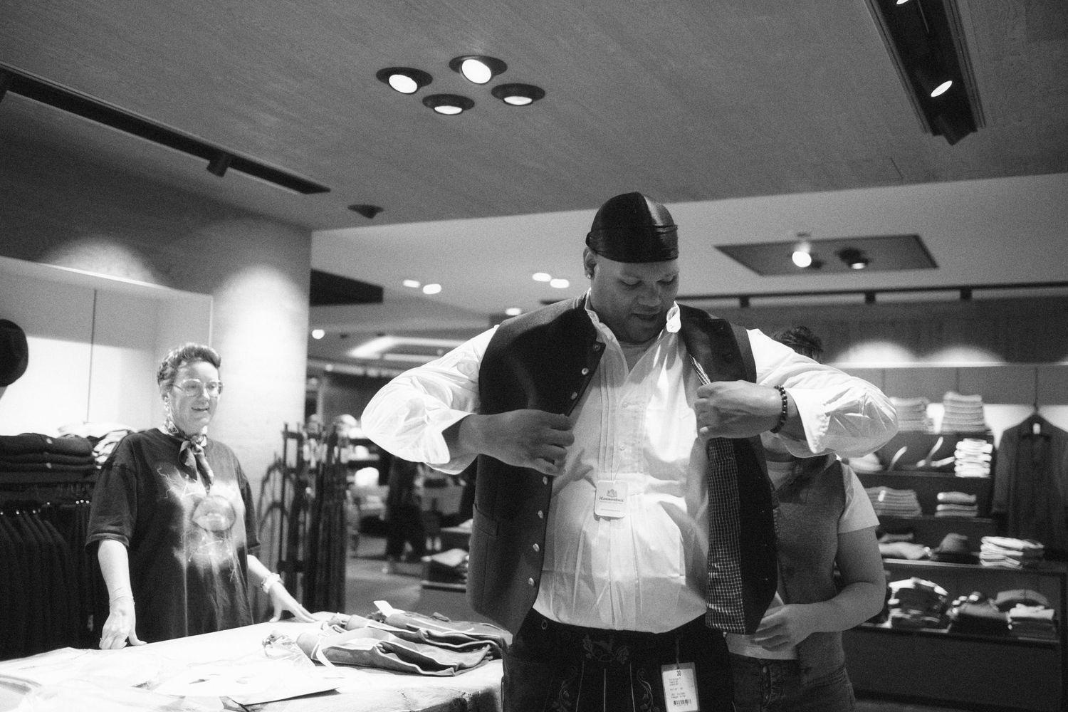 Man trying on jacket in clothing store, black and white photo showing retail shopping experience with staff member in background.