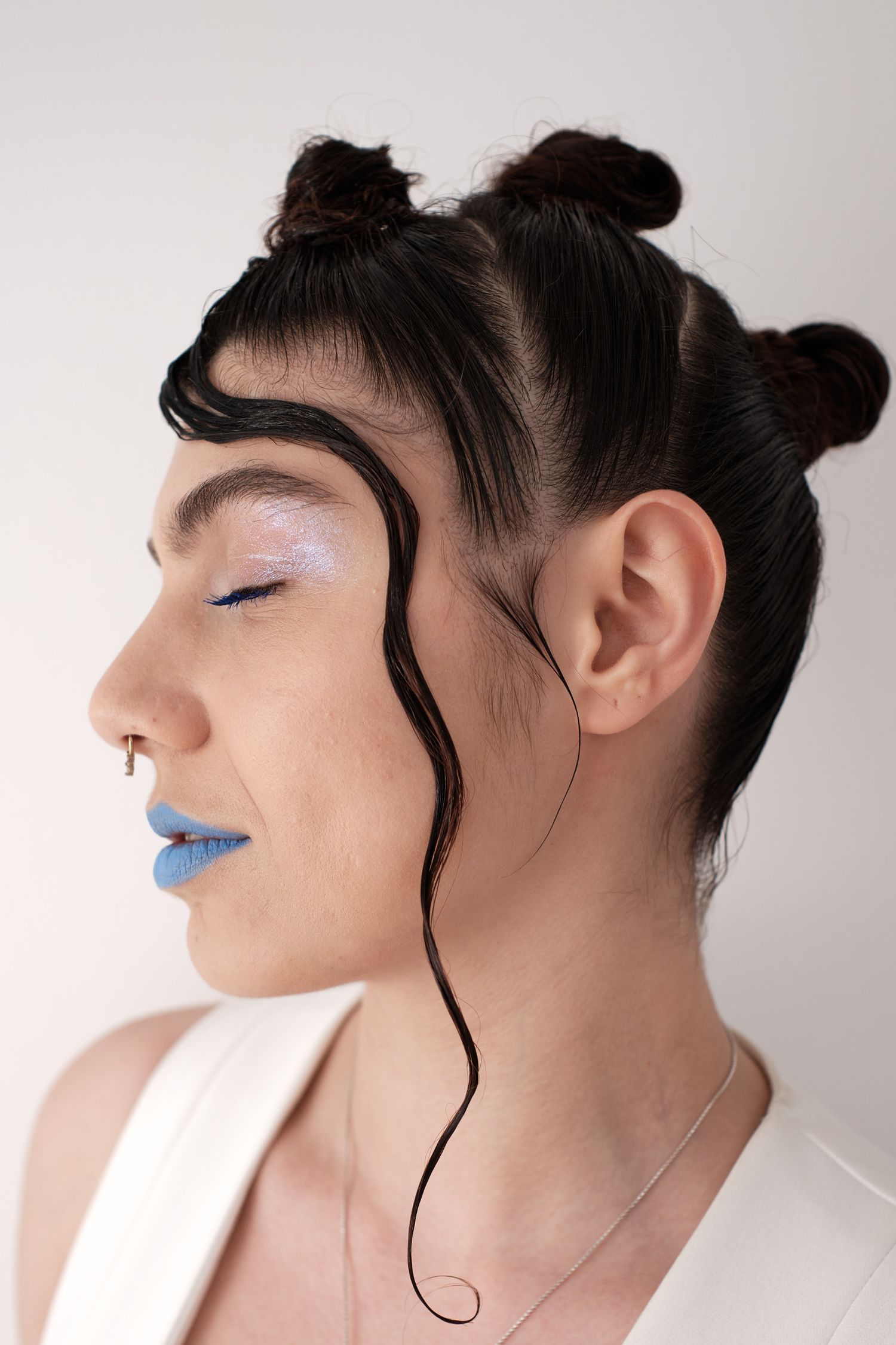 Woman with blue lips, nose ring, double bun hairstyle, and artistic wavy hair tendrils, wearing white top.