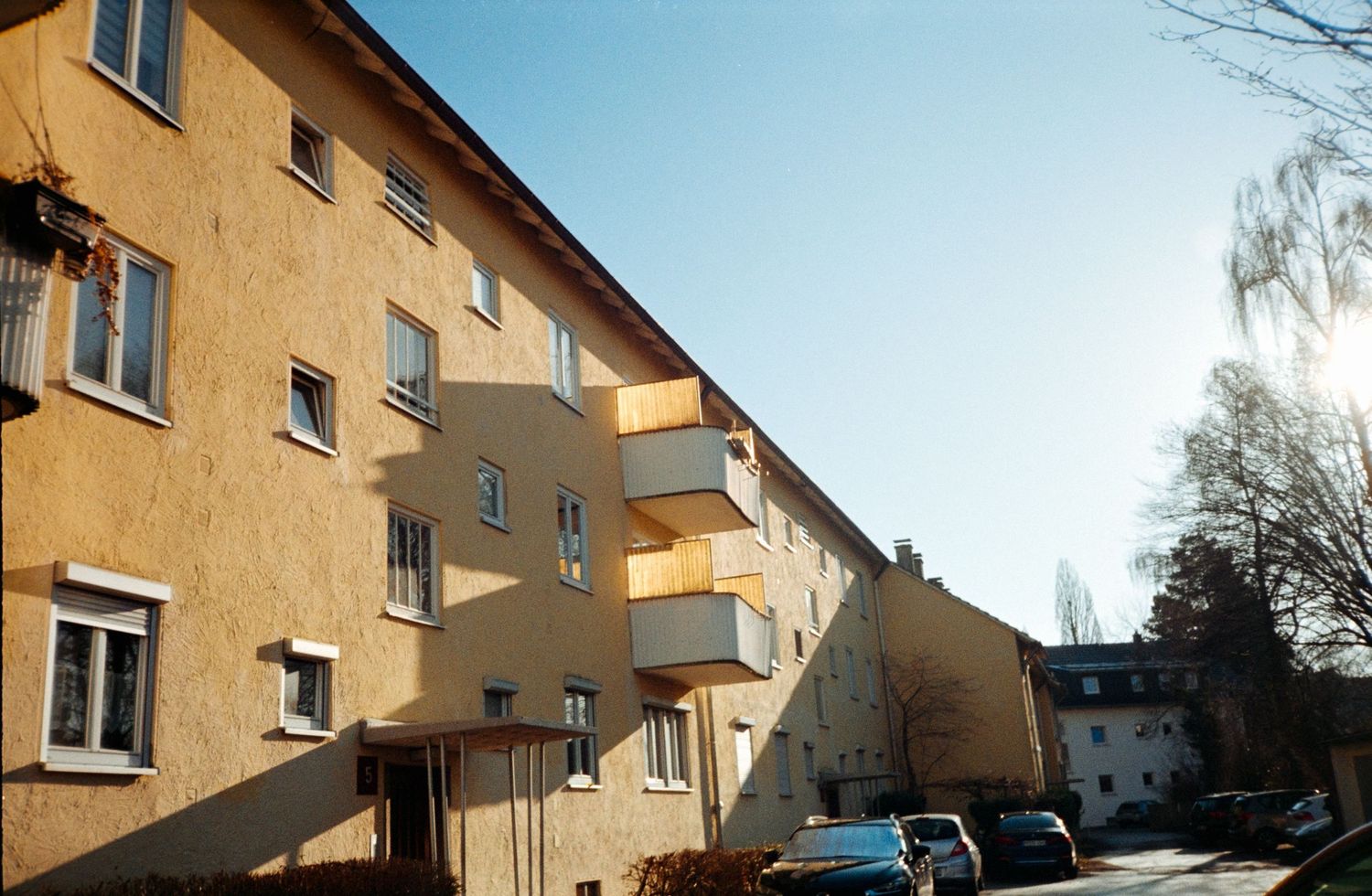 Yellow multi-story residential apartment building with balconies and bare trees on a sunny winter day.