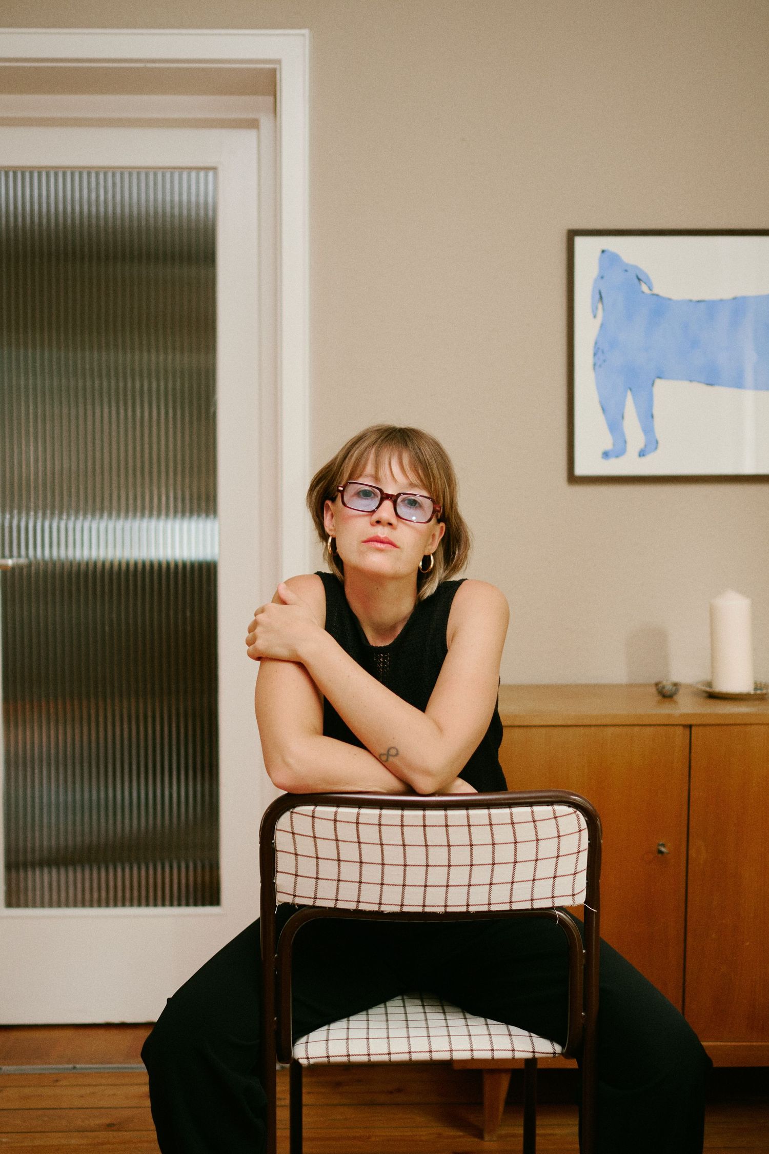 Woman in glasses and black outfit sits on a vintage chair in a mid-century modern room with artwork on the wall.