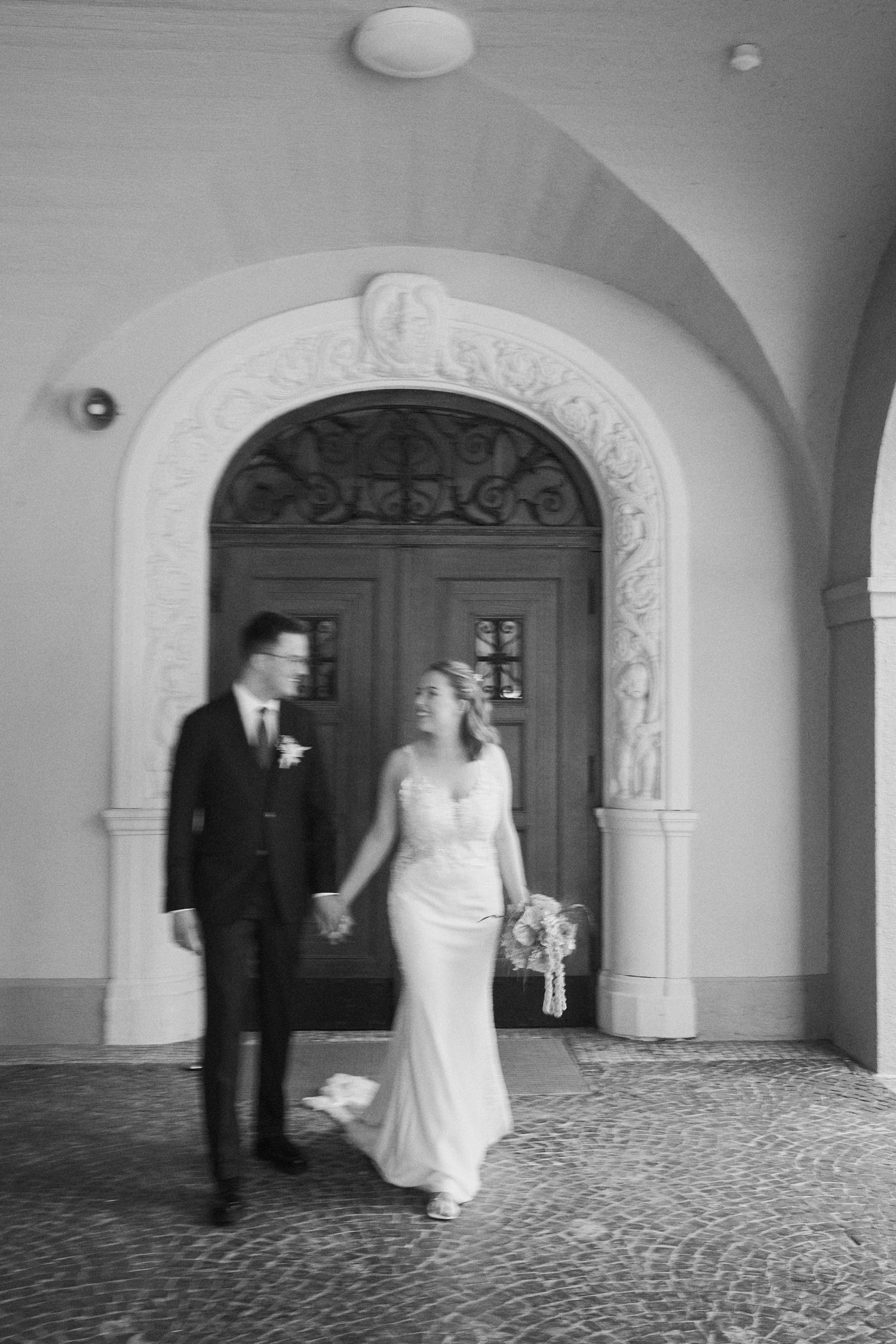 Black and white photo of a bride and groom holding hands walking under an ornate arch doorway.