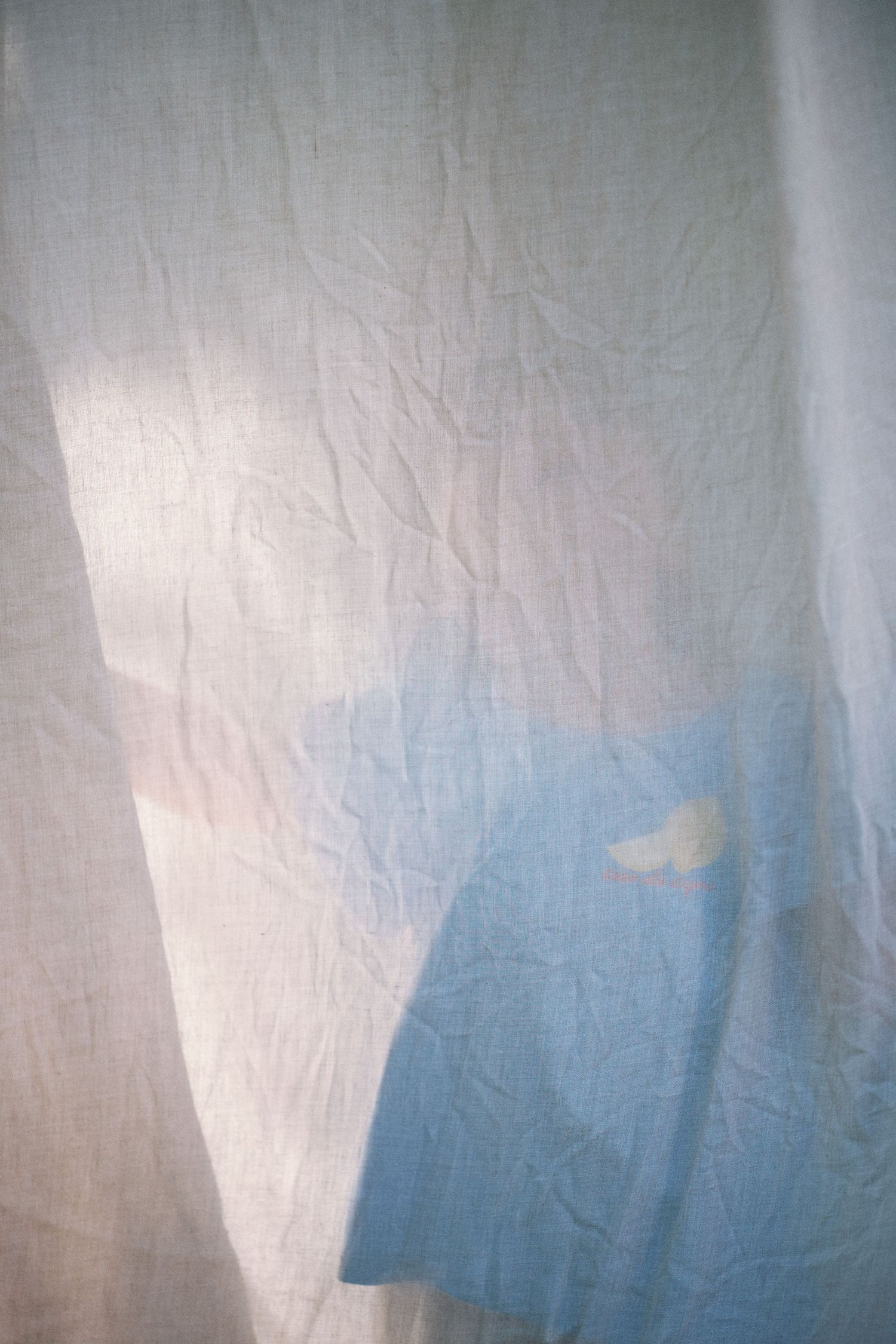 Sheer white linen curtain with soft wrinkles, backlit by natural light revealing a blue fabric shadow behind.