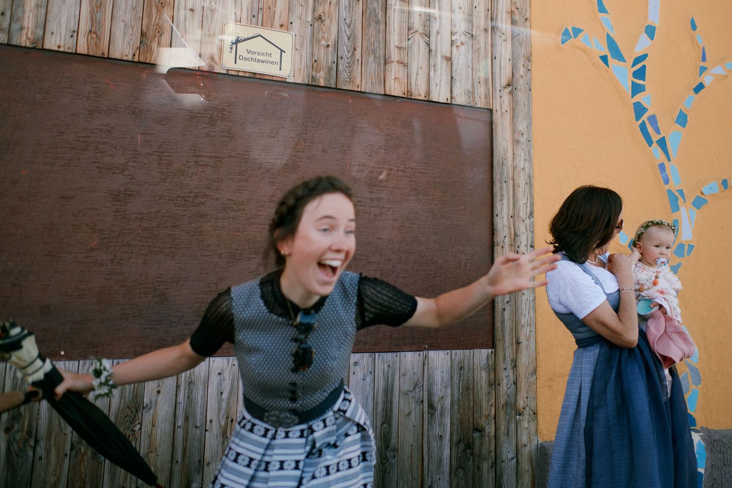 A joyful woman in costume spreads her arms wide while another woman holds a toddler nearby outdoors.