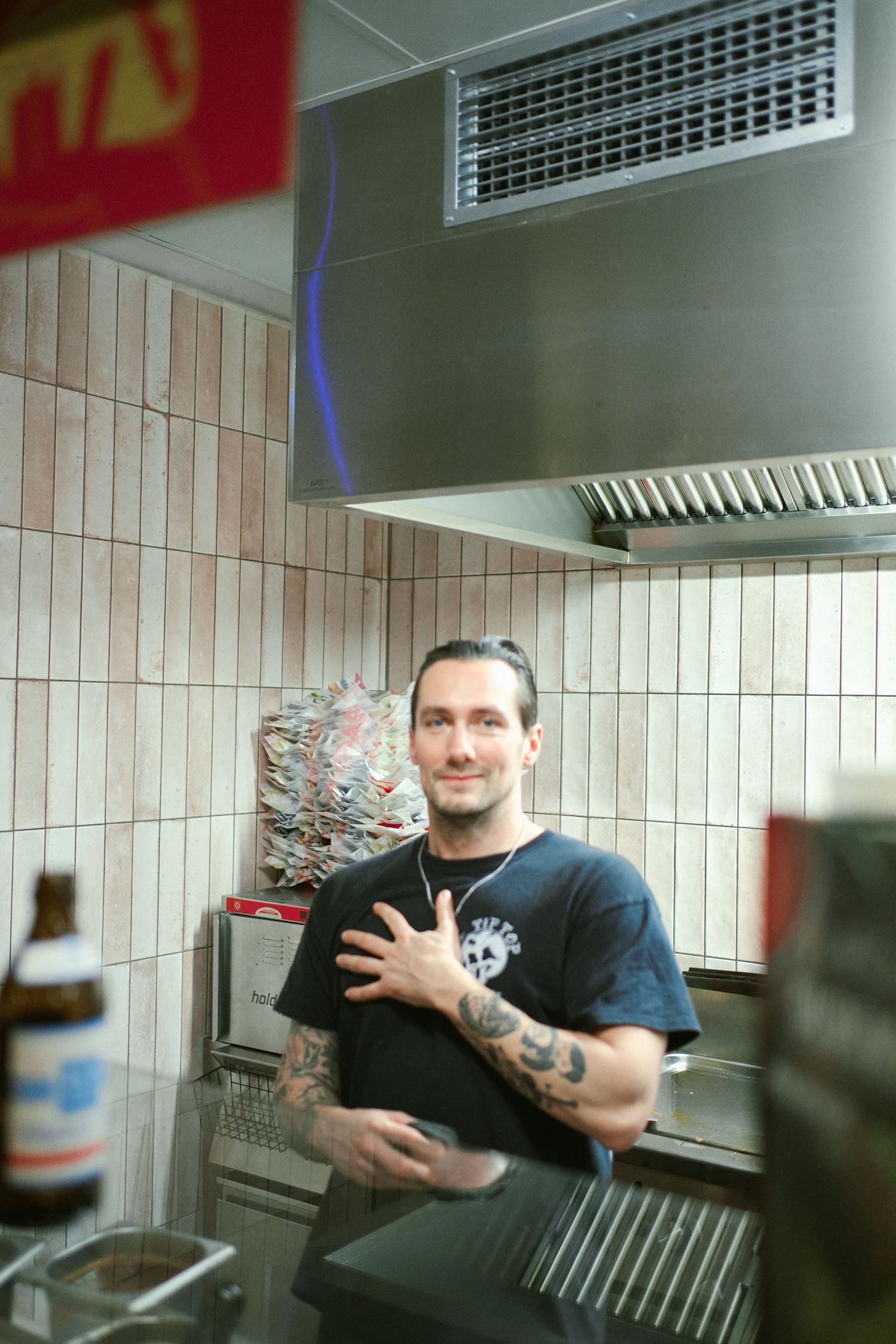 Tattooed male chef smiling in a commercial kitchen with tiled walls and stainless steel hood vent.