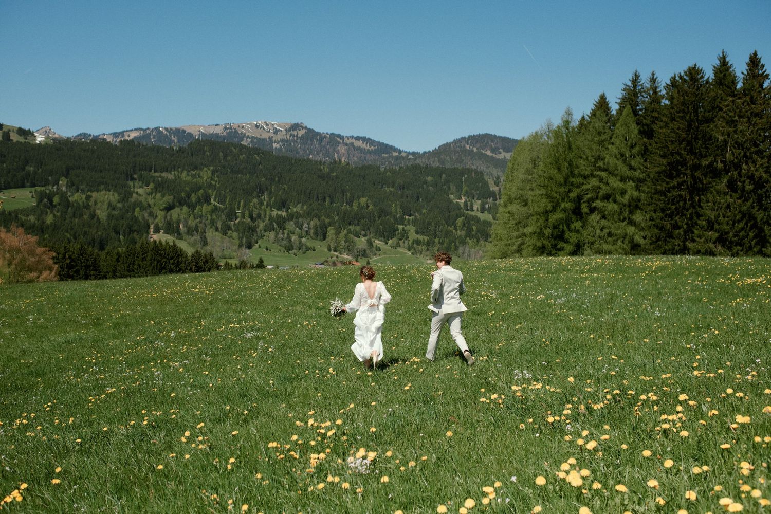 Couple in white attire running through a lush green meadow with yellow wildflowers and mountain backdrop.