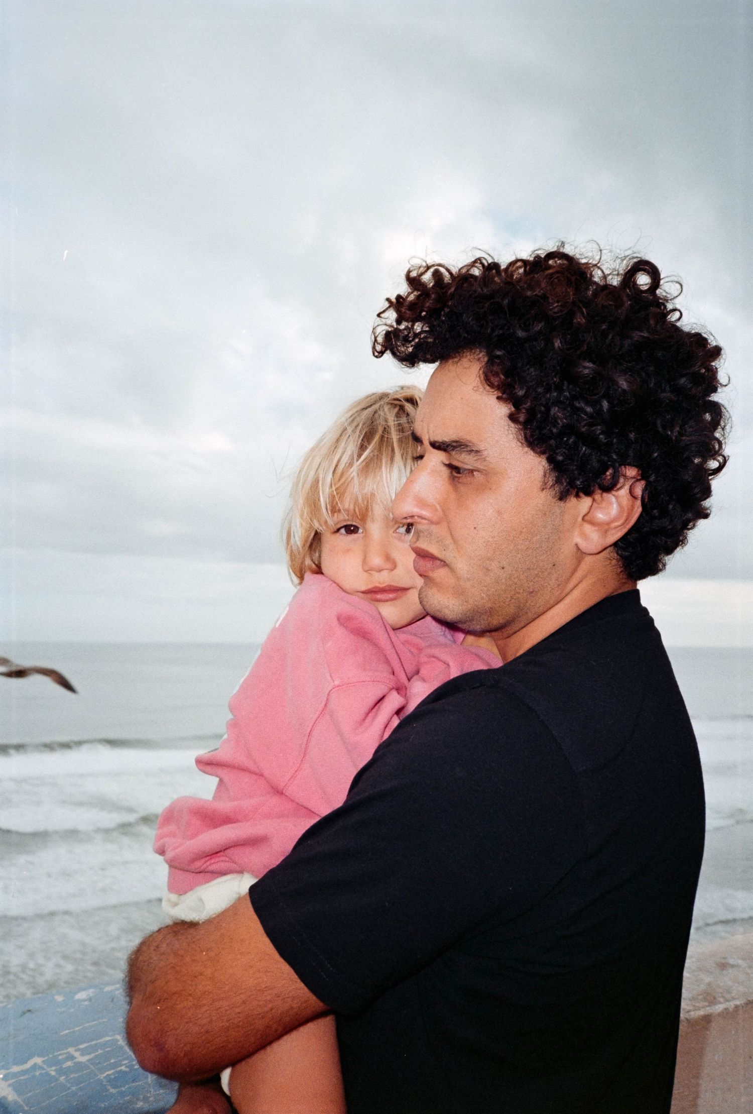 A curly-haired man holds a young blonde child in a pink outfit near a cloudy ocean beach.