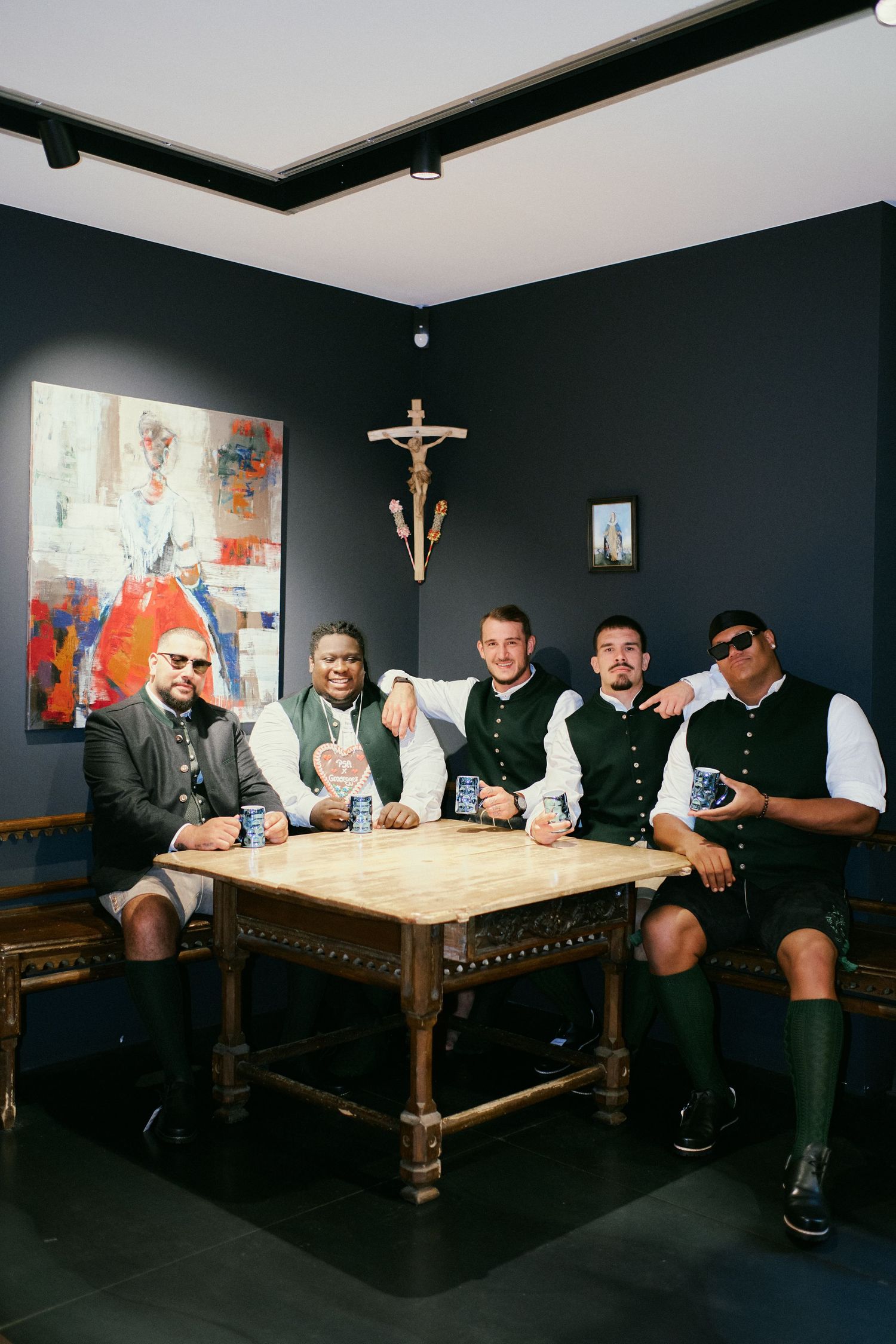 Five men in traditional Bavarian attire pose together at a wooden table holding drinks in a dark-walled room.