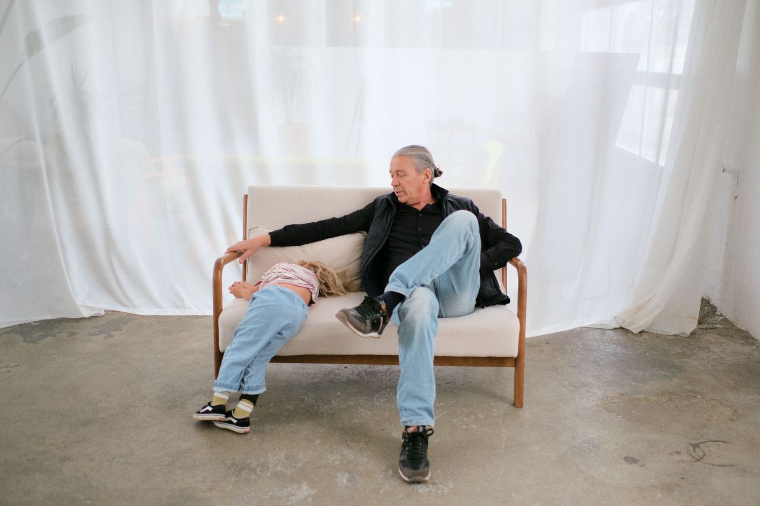 Two people sitting together on a wooden bench against white curtains on a concrete floor.