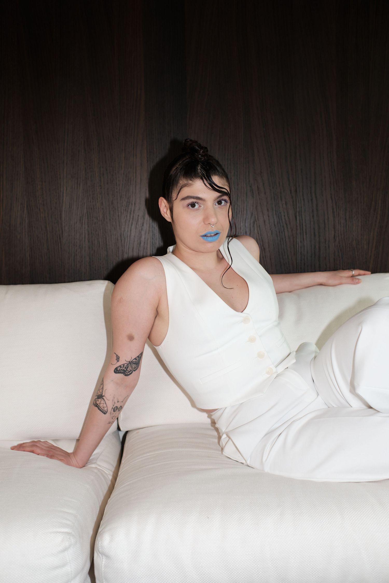 Person in white outfit with blue lipstick and arm tattoo sitting on white sofa against dark wood wall.