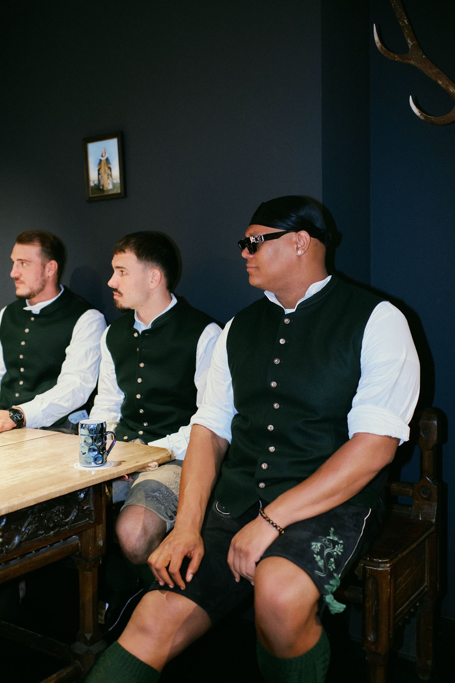 Three men in traditional Bavarian attire with dark vests and white shirts seated in a beer hall.