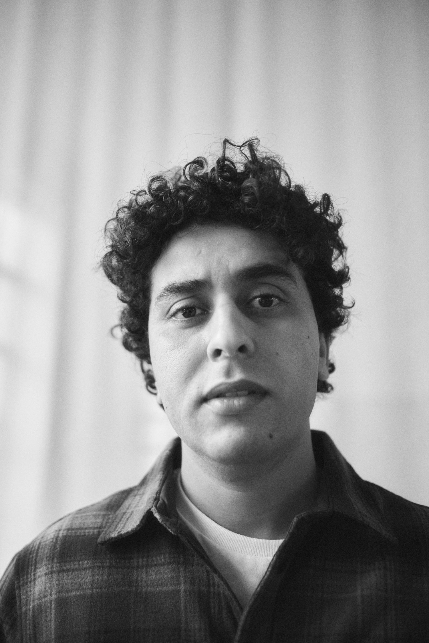 Black and white portrait of a young man with curly hair wearing a plaid shirt, looking directly at the camera.