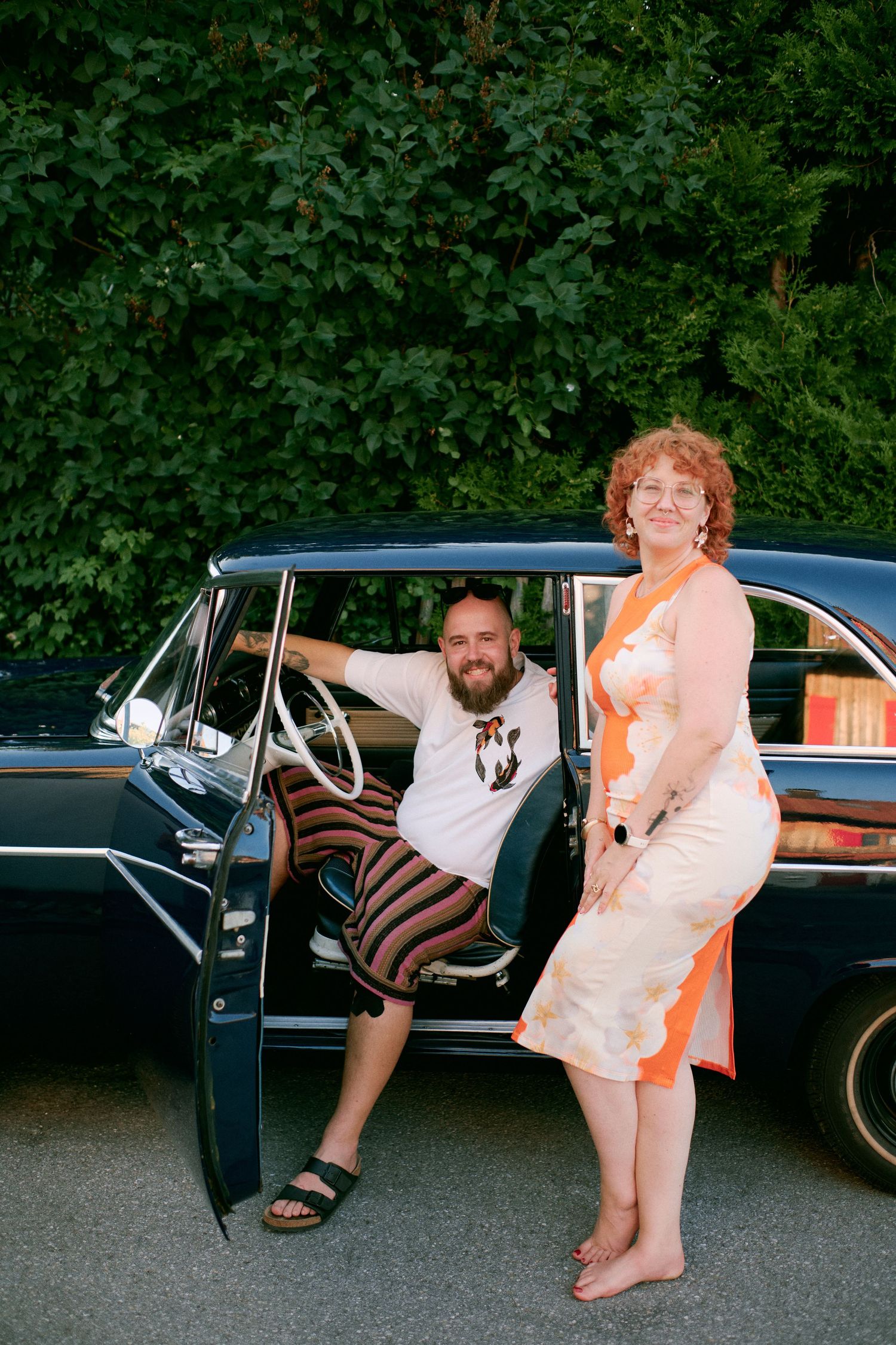 A bearded man and a curly red-haired woman pose beside a vintage dark blue classic car outdoors.