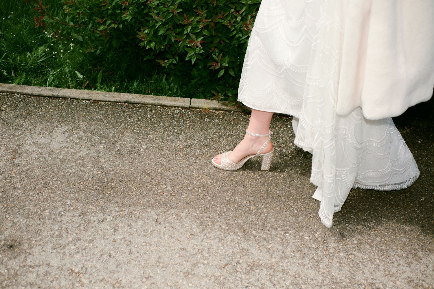 Close-up of a bride wearing a white wedding dress and blush pink heeled shoes on a gravel path.