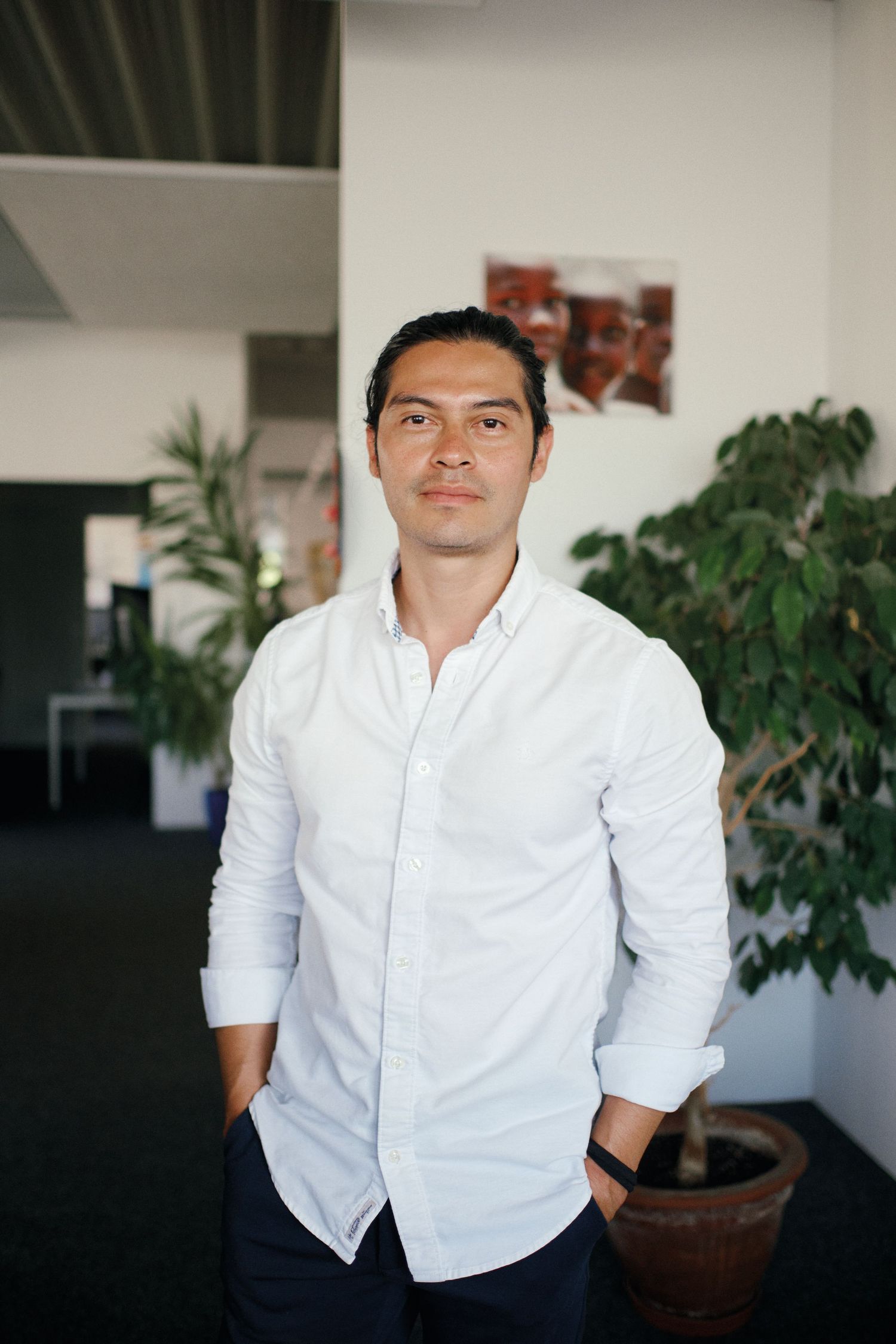 A confident young man in a white button-up shirt stands in a modern office with green plants.