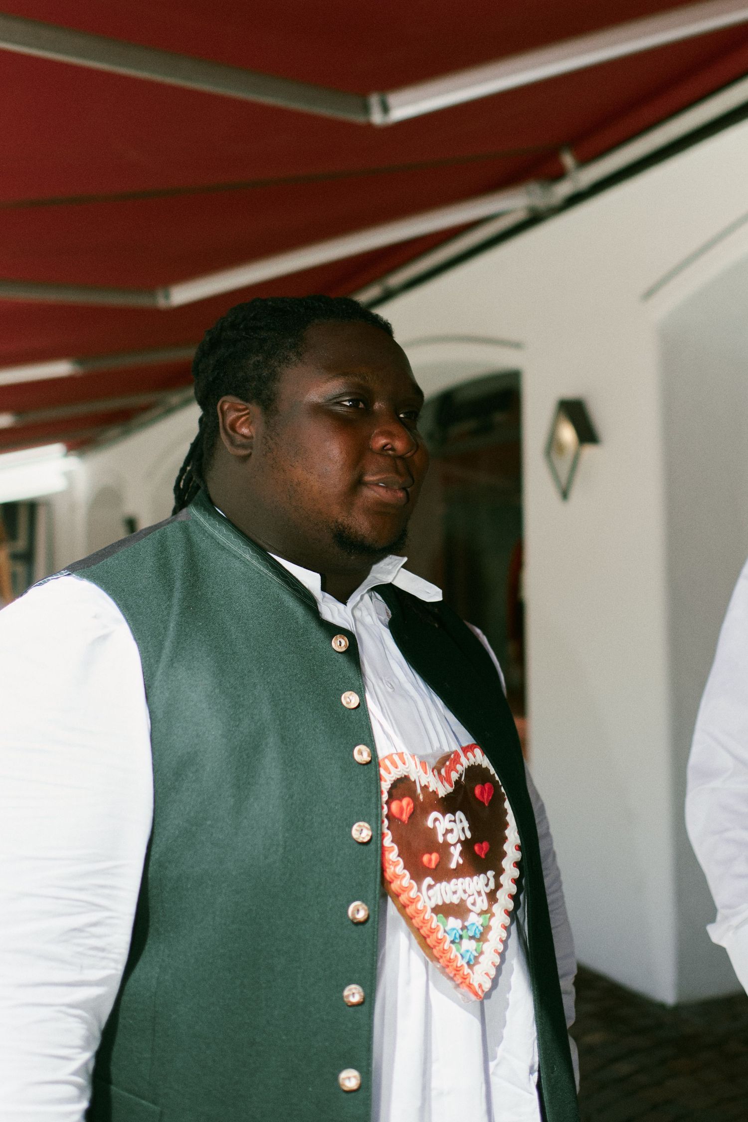 Man in traditional Bavarian vest with decorative gingerbread heart necklace standing outdoors.