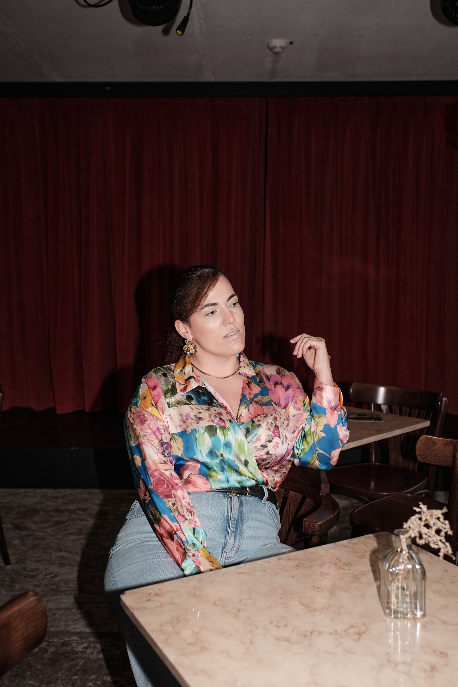 Woman in colorful floral blouse and jeans seated at marble table in dimly lit lounge with red curtains.