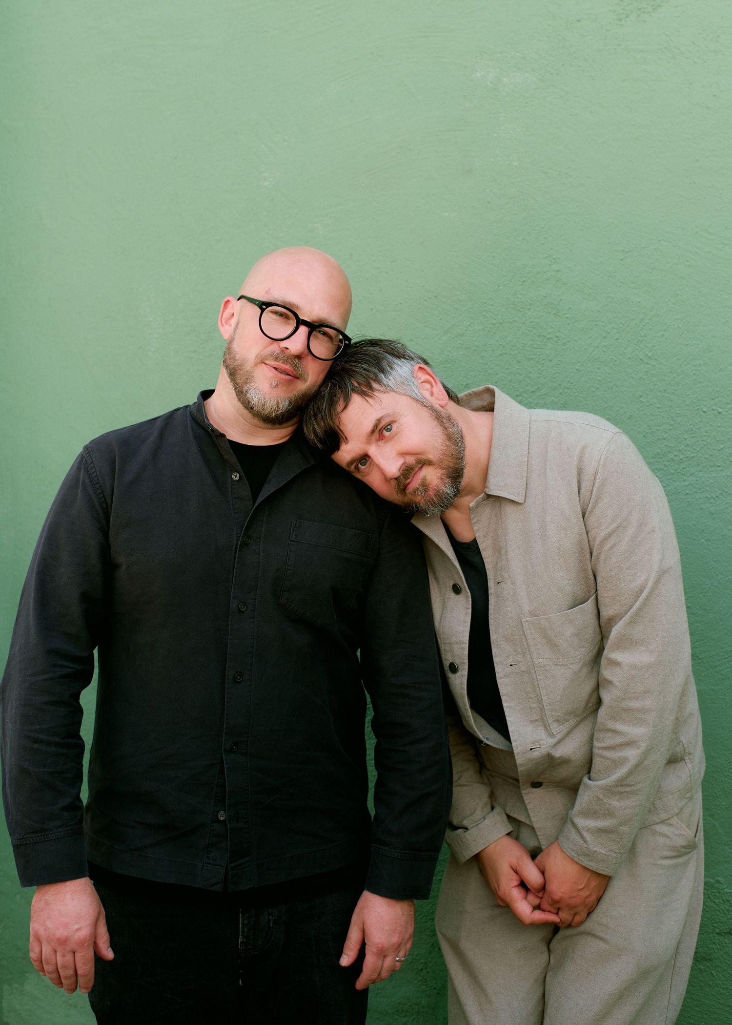 Two bearded men pose warmly against a green wall, one bald with glasses, the other leaning his head on his shoulder.