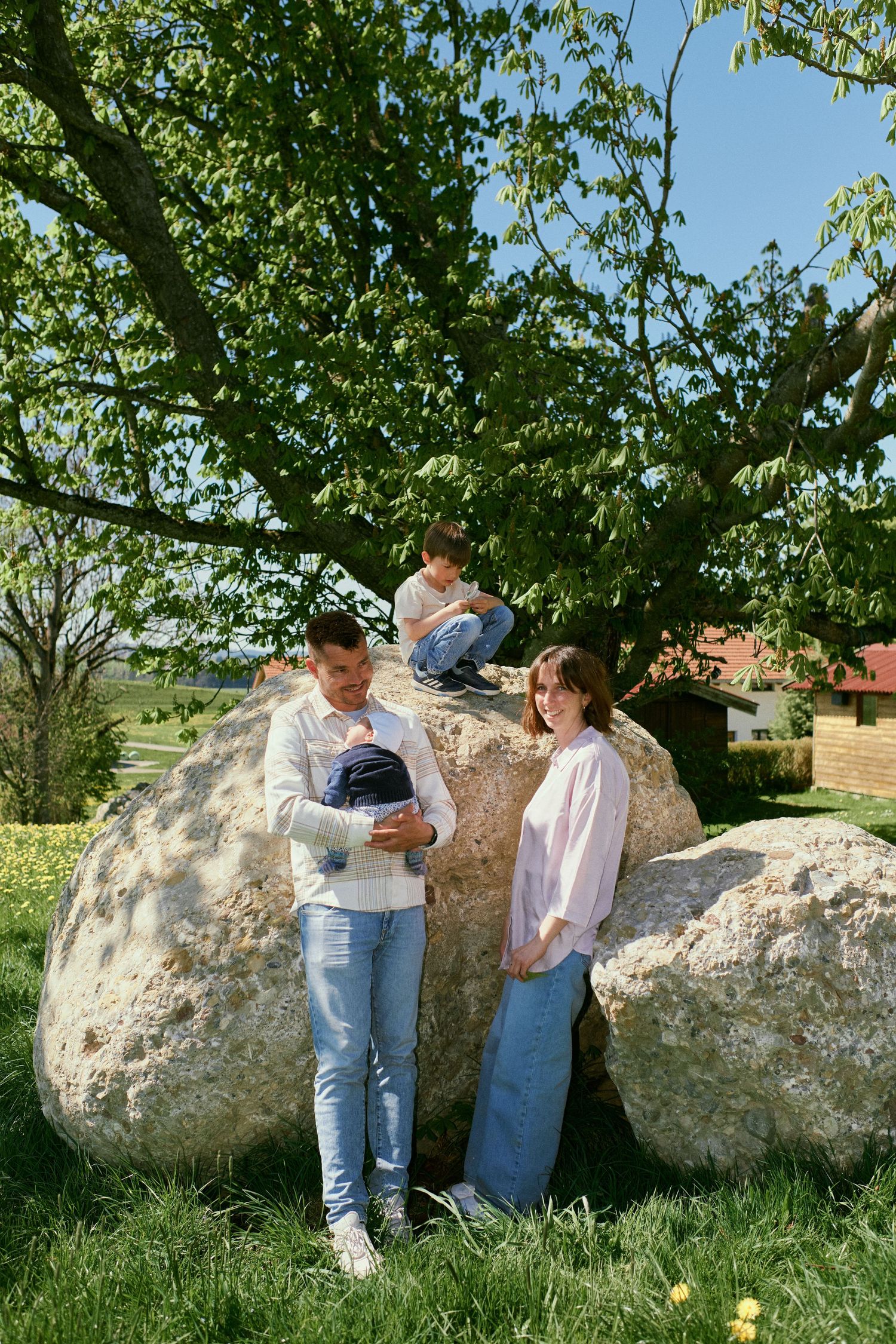Family of four enjoying sunny day outdoors near large boulders and green tree, parents holding baby while child sits on rock.