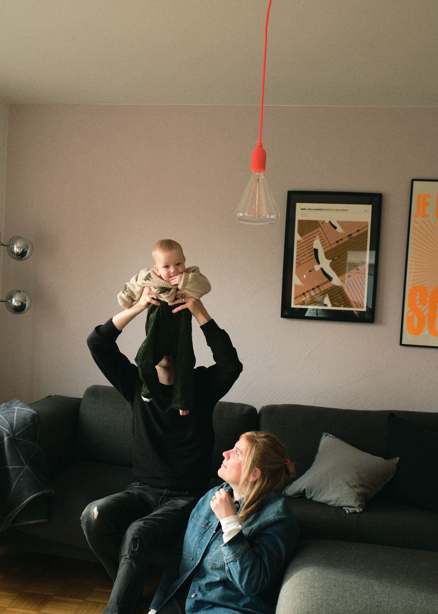 A parent lifts a baby above their head while another adult relaxes on a gray sofa in a modern living room.