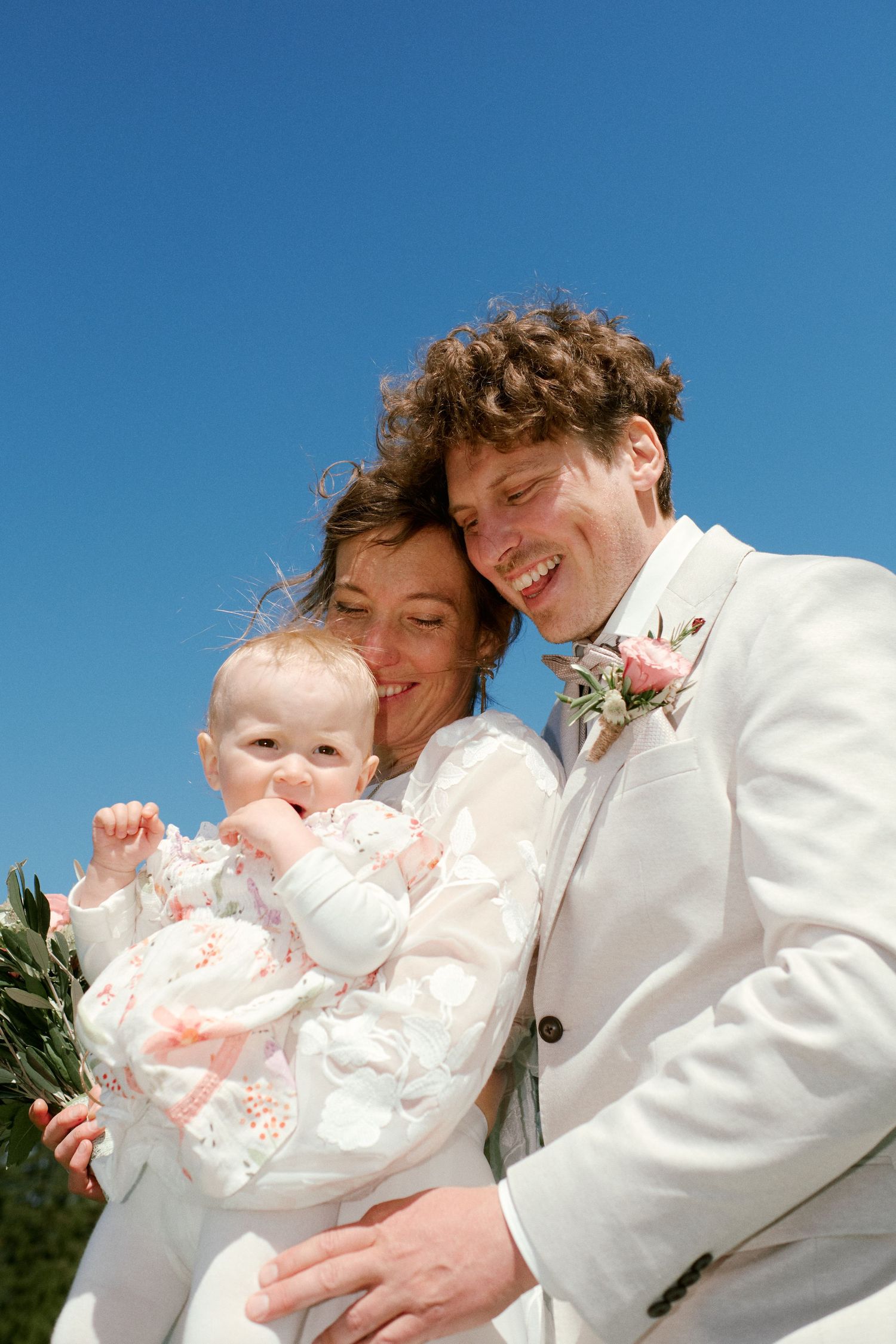 Happy newlywed couple in white attire smiling while holding their baby girl under a clear blue sky.