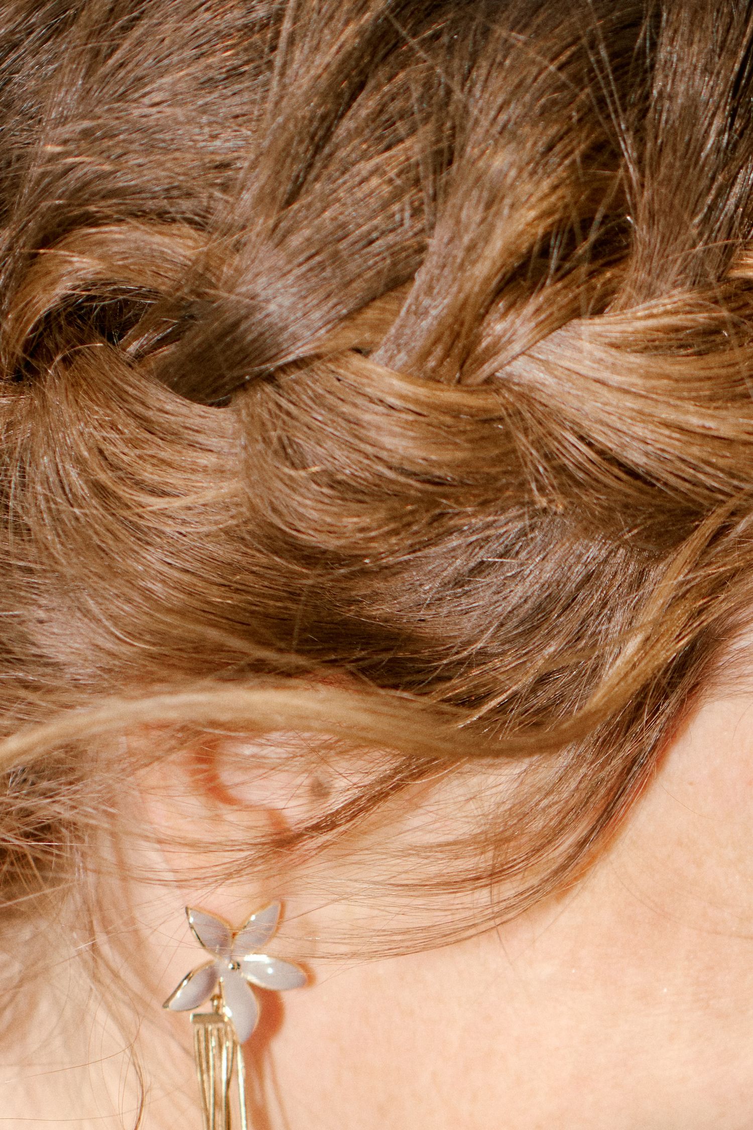 Close-up of a woman's braided auburn hair with a delicate gold flower earring visible near her ear.