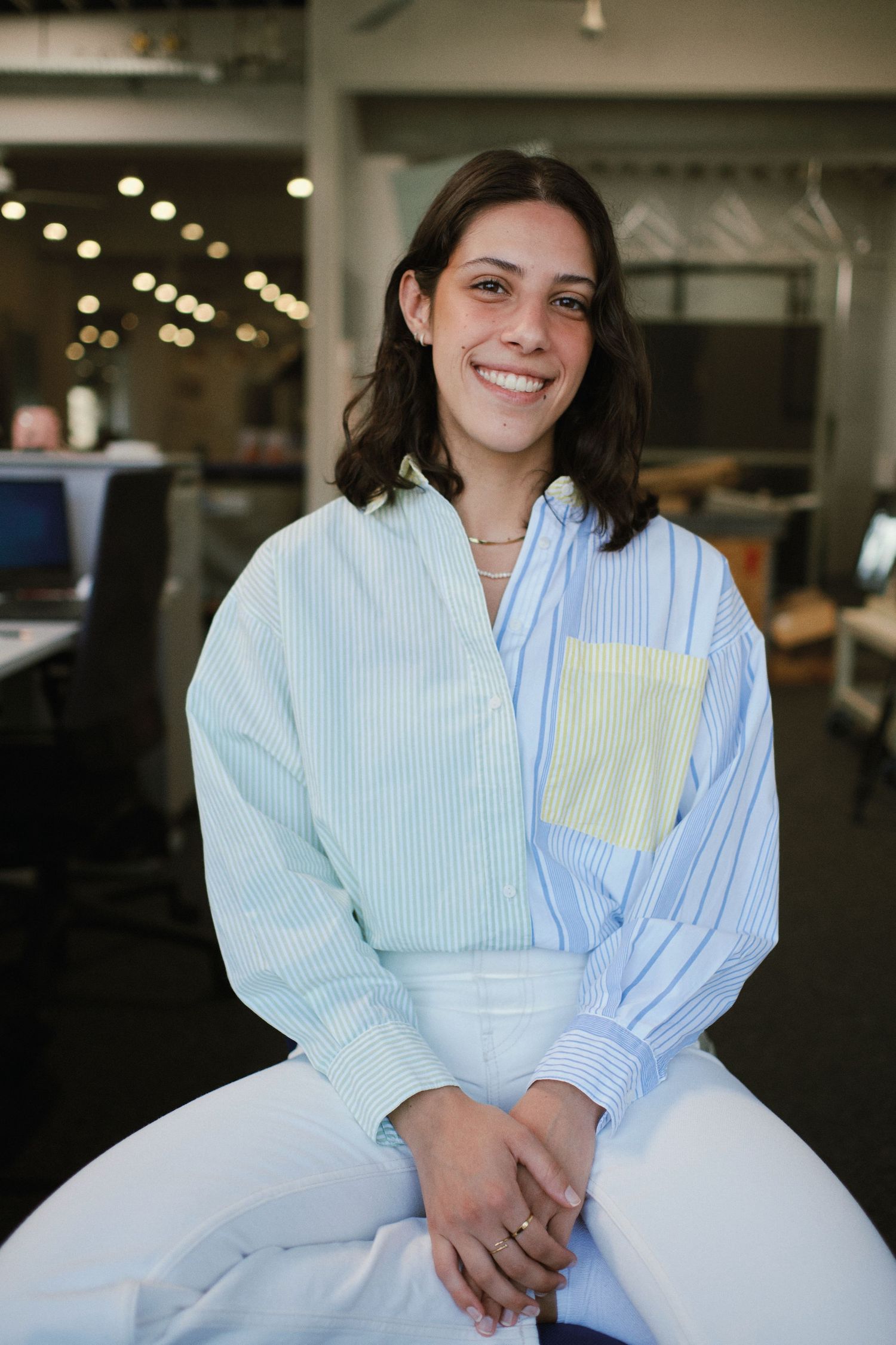 Smiling young woman in striped blue shirt sitting in modern office environment.