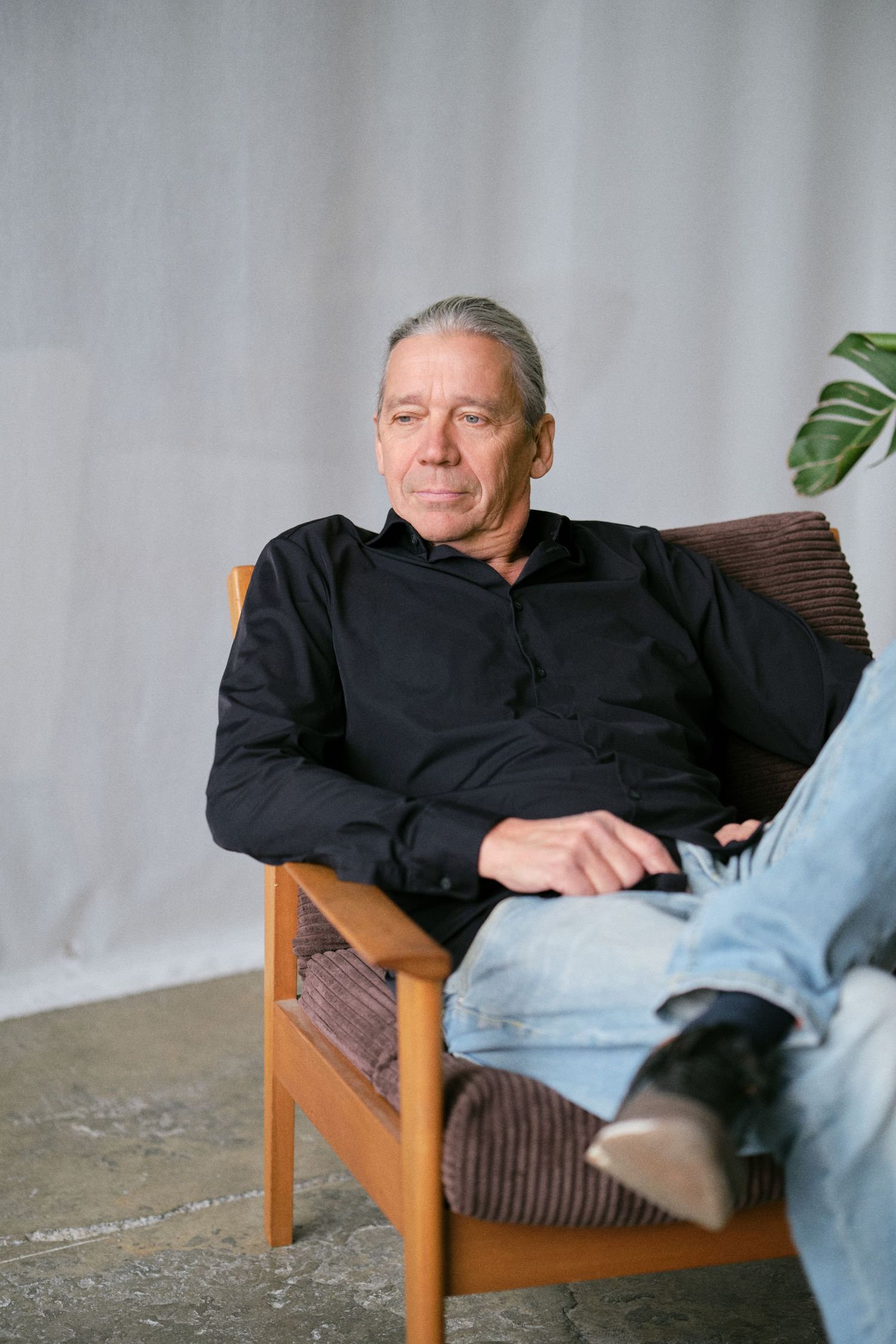 Middle-aged man in black shirt and jeans relaxing in wooden armchair against light grey backdrop.