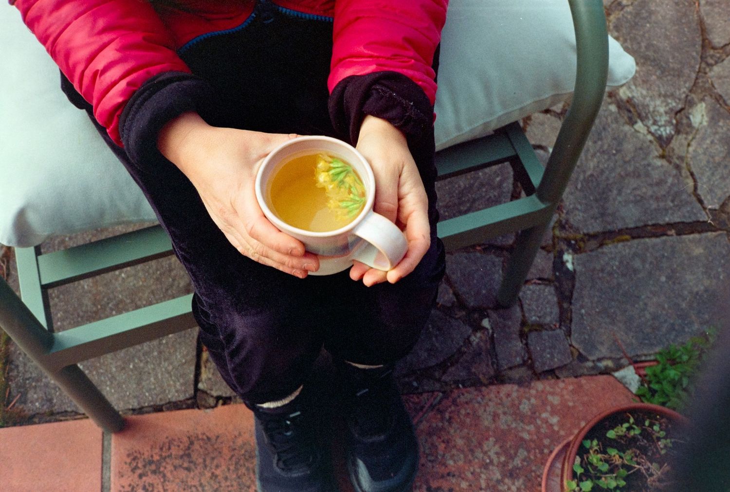 Person in red jacket holding a white mug of golden tea while seated outdoors on a green chair.