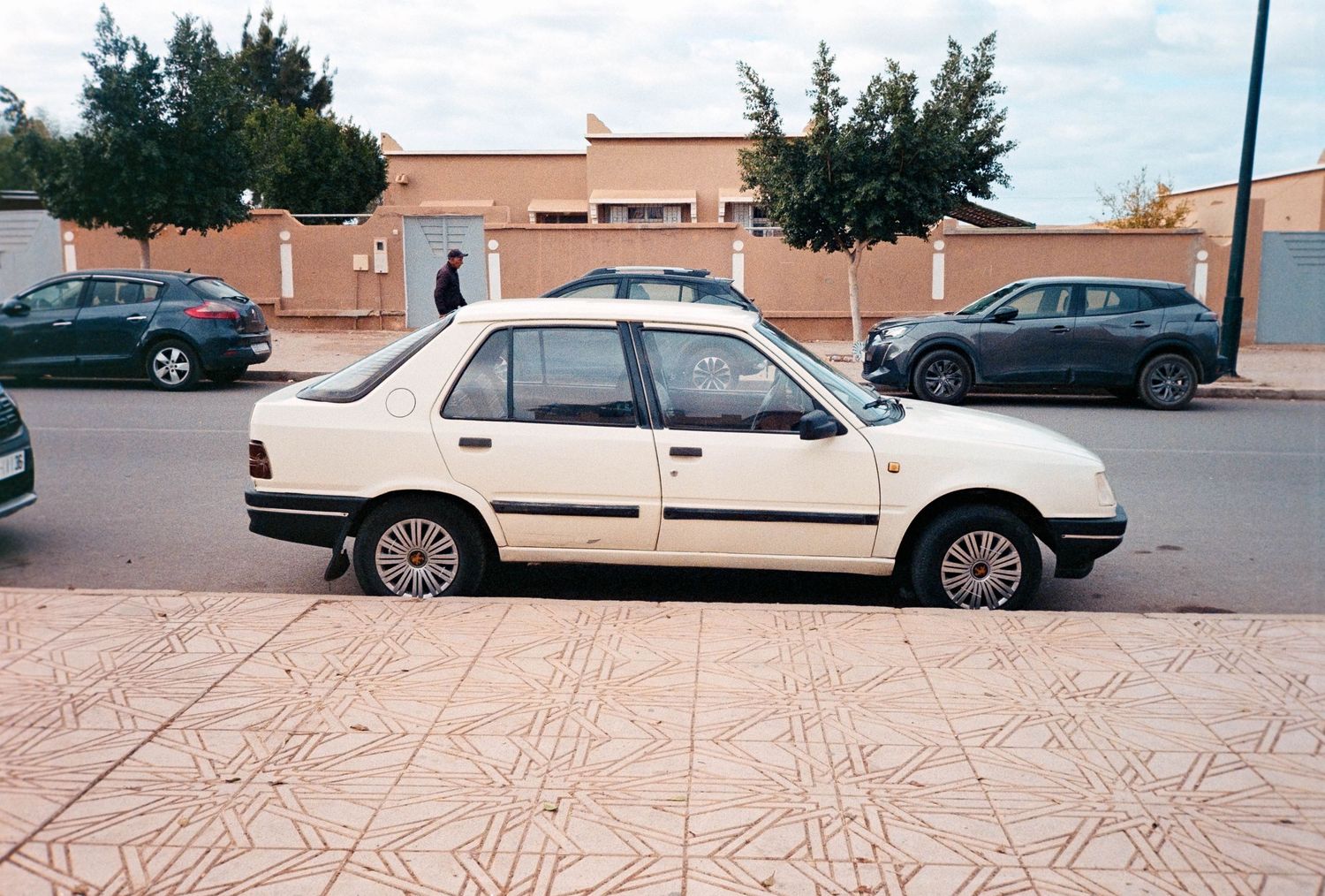 White sedan car parked on a tiled sidewalk in a sunny residential street with other vehicles in the background.