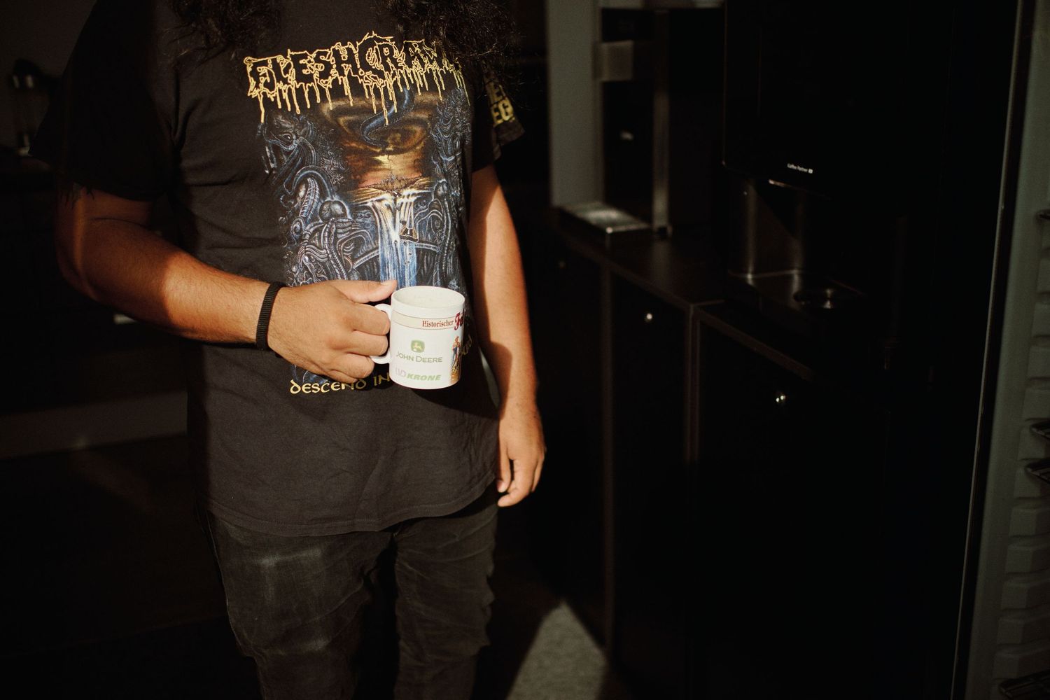 Person in black metal band t-shirt holding a white coffee cup in a dimly lit room.