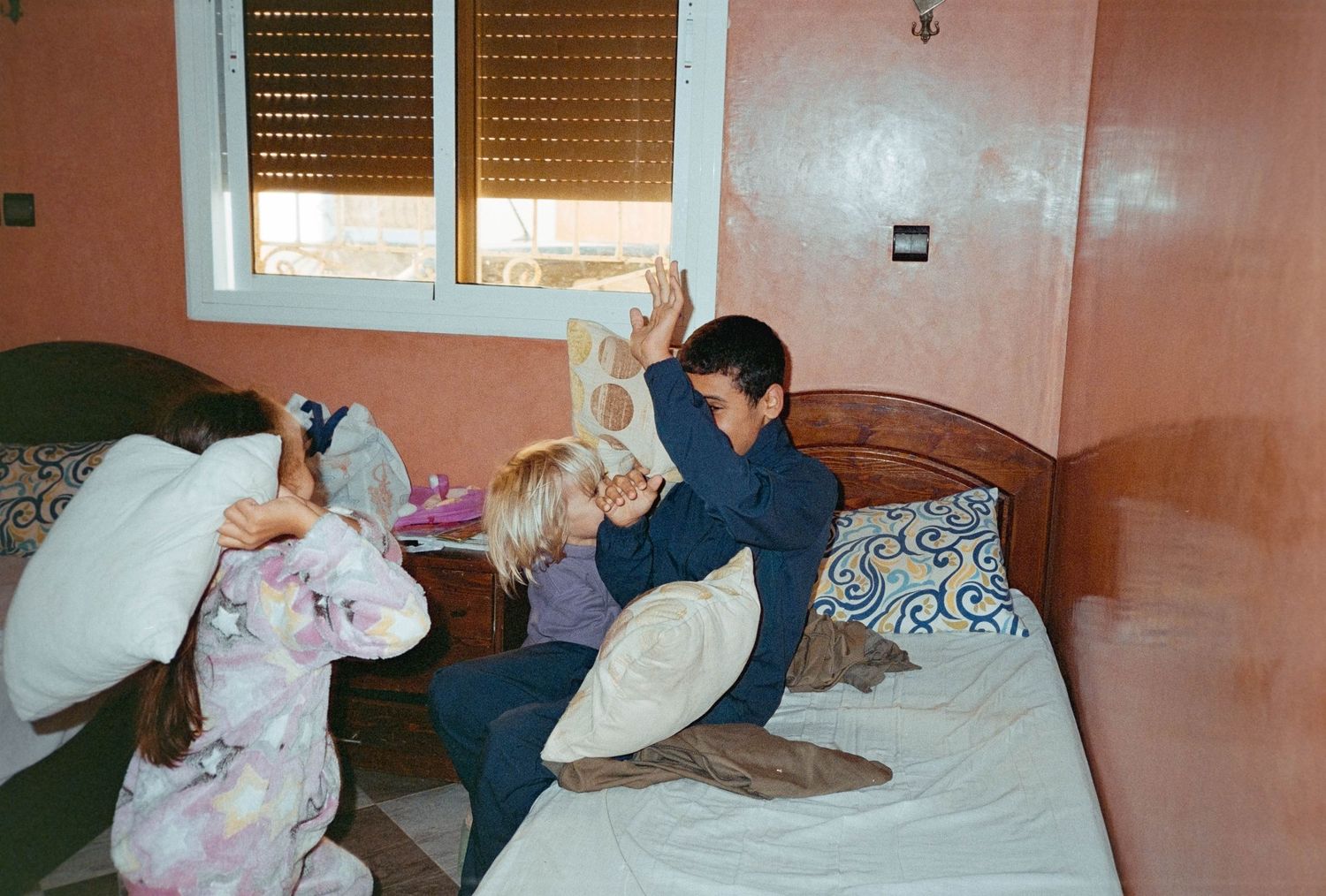 Three children having a pillow fight on a bed in a cozy bedroom with pink walls and a window.