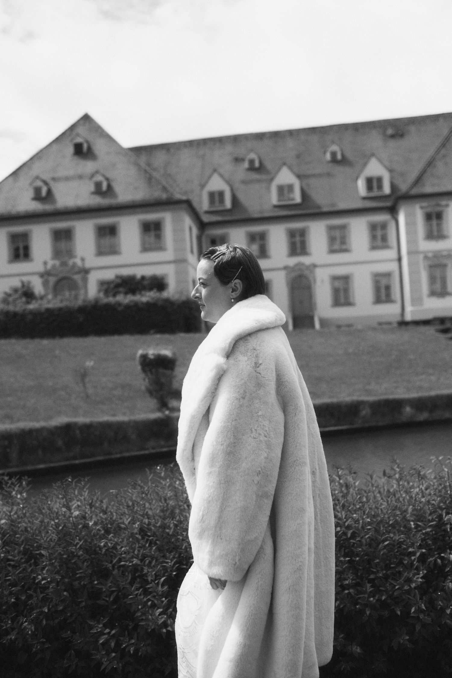 Black and white photo of a woman in a long fur coat standing before a historic European manor house.
