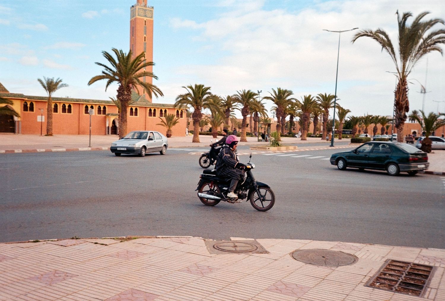Motorcyclist riding through a sunny Moroccan town square with palm trees, parked cars, and a mosque minaret in the background.