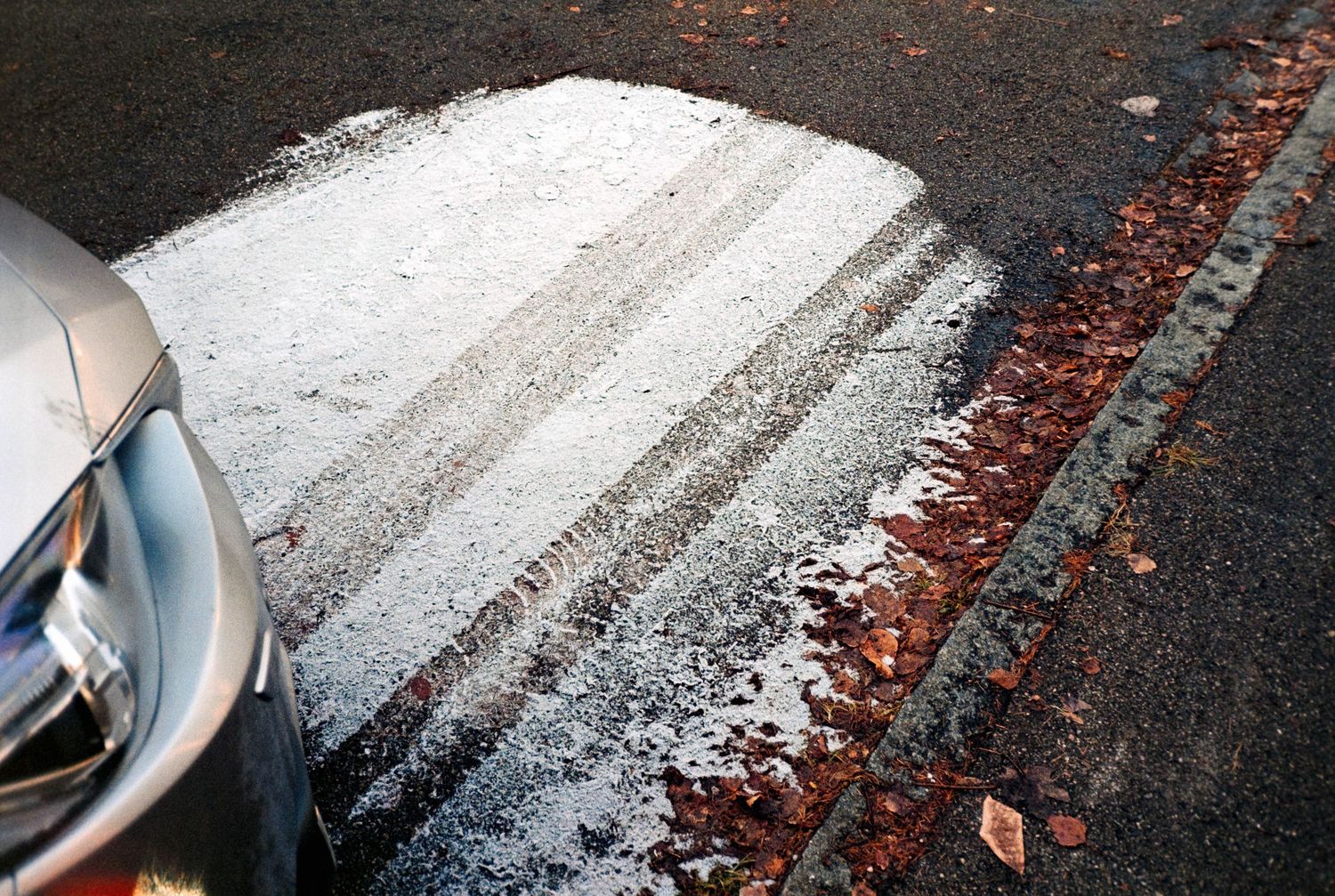 Icy road surface with tire tracks in frost near fallen autumn leaves and a parked car.