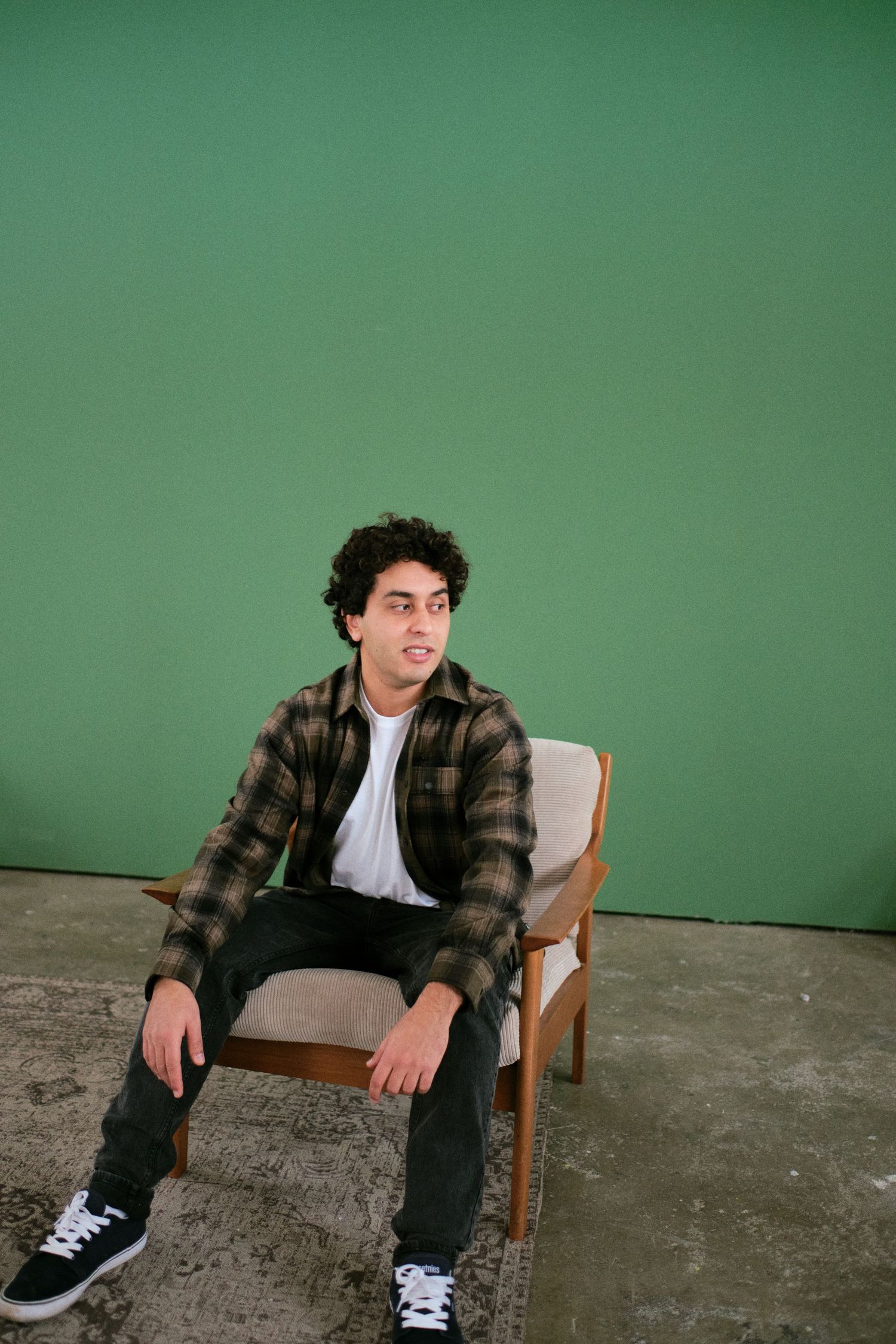 Young man in plaid shirt and black jeans sitting on a wooden chair against a green background.