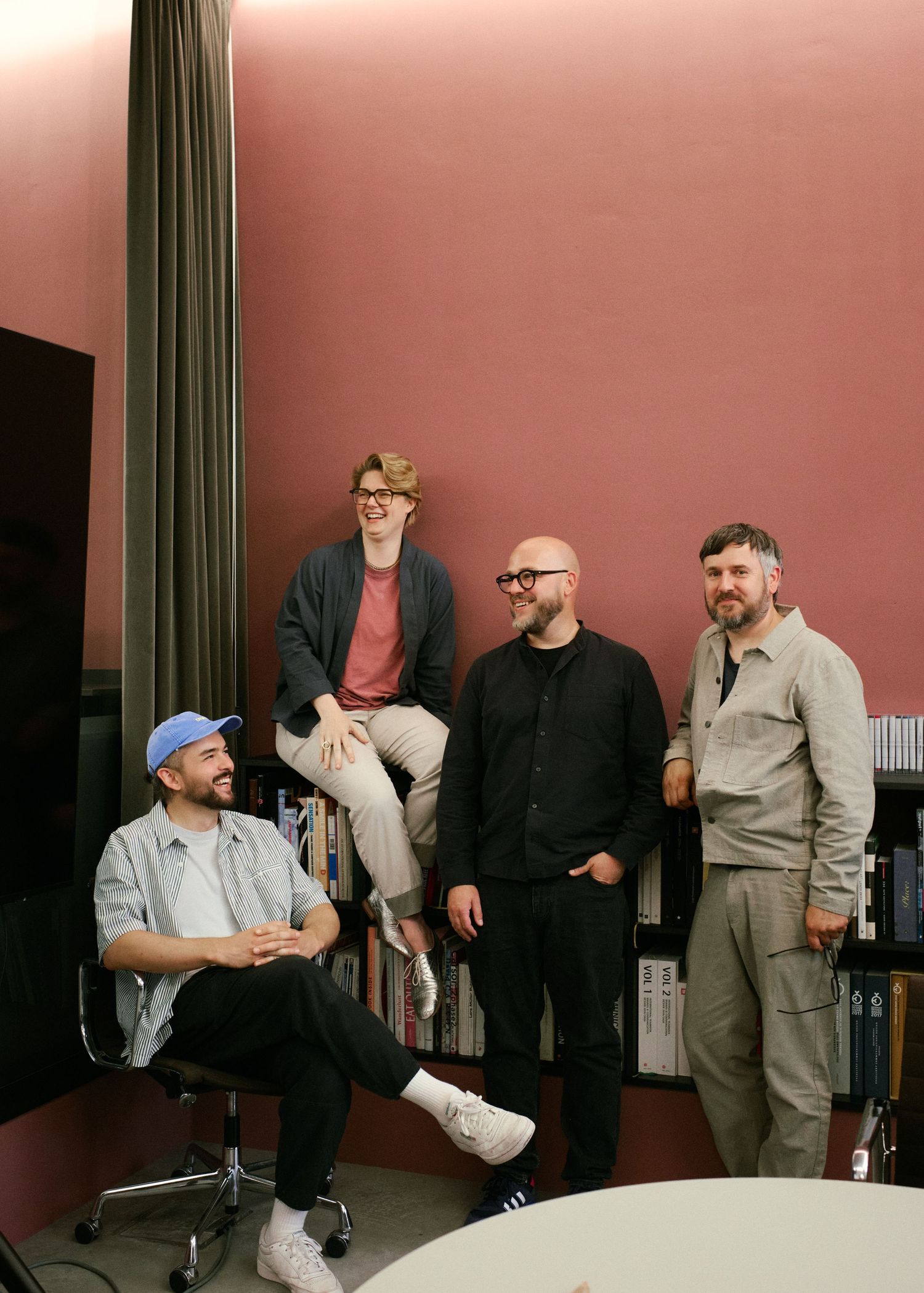 Four men posing together in a stylish pink-walled room, smiling and relaxed in casual and smart outfits.