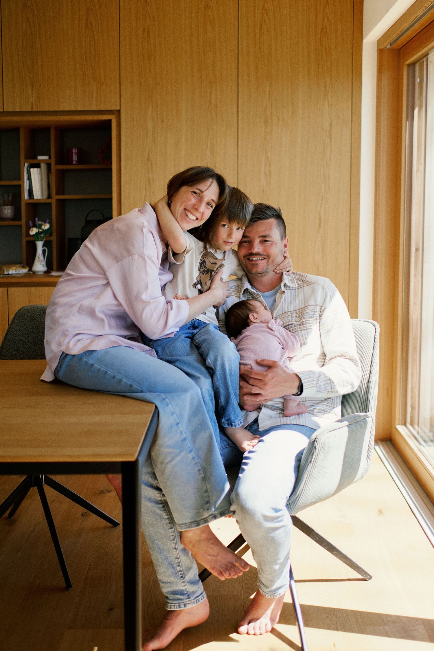 Happy family portrait with parents and two children, including a newborn baby, sitting together in a modern wooden interior.