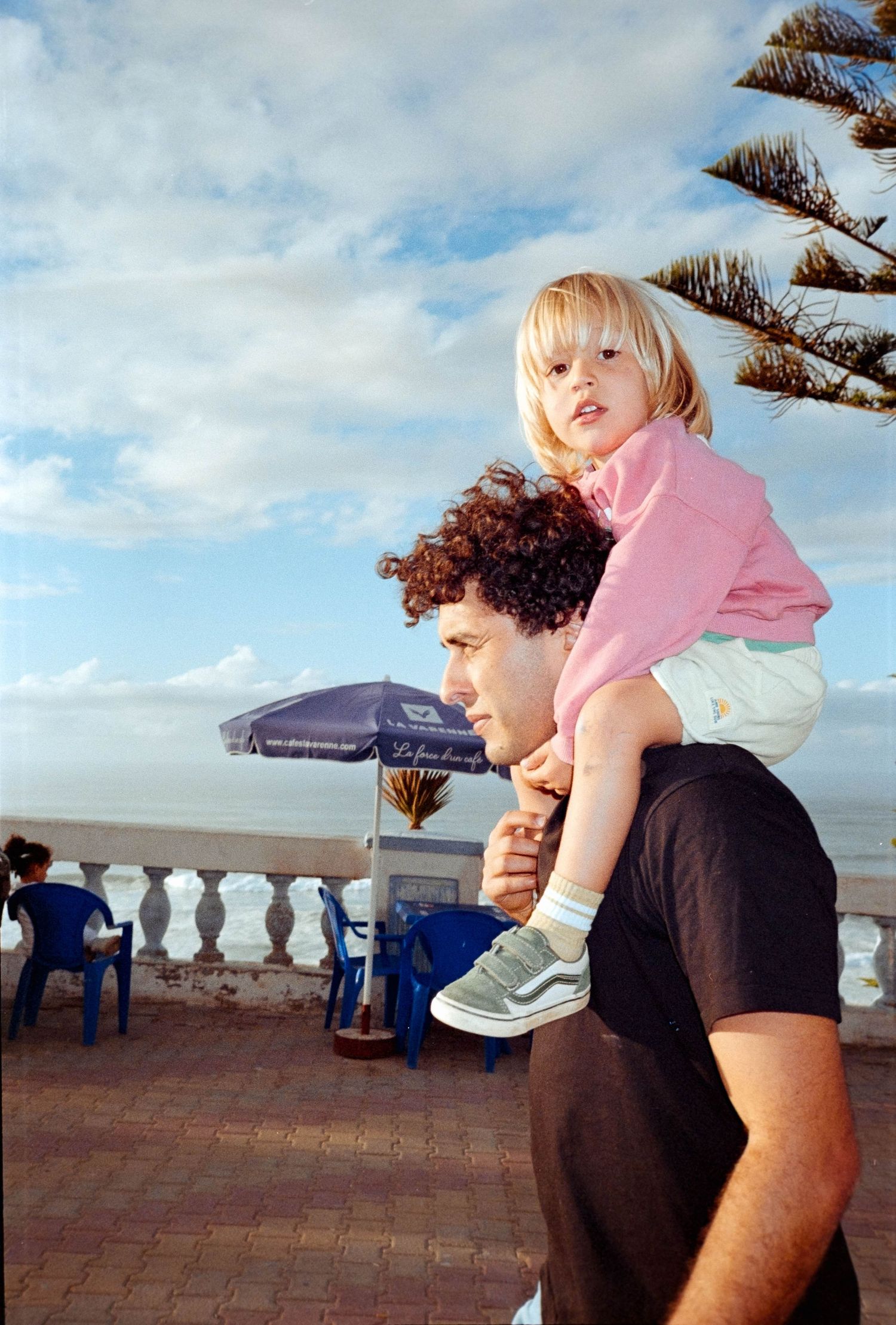 A curly-haired man carries a young blonde girl in a pink shirt on his shoulders near a seaside terrace.